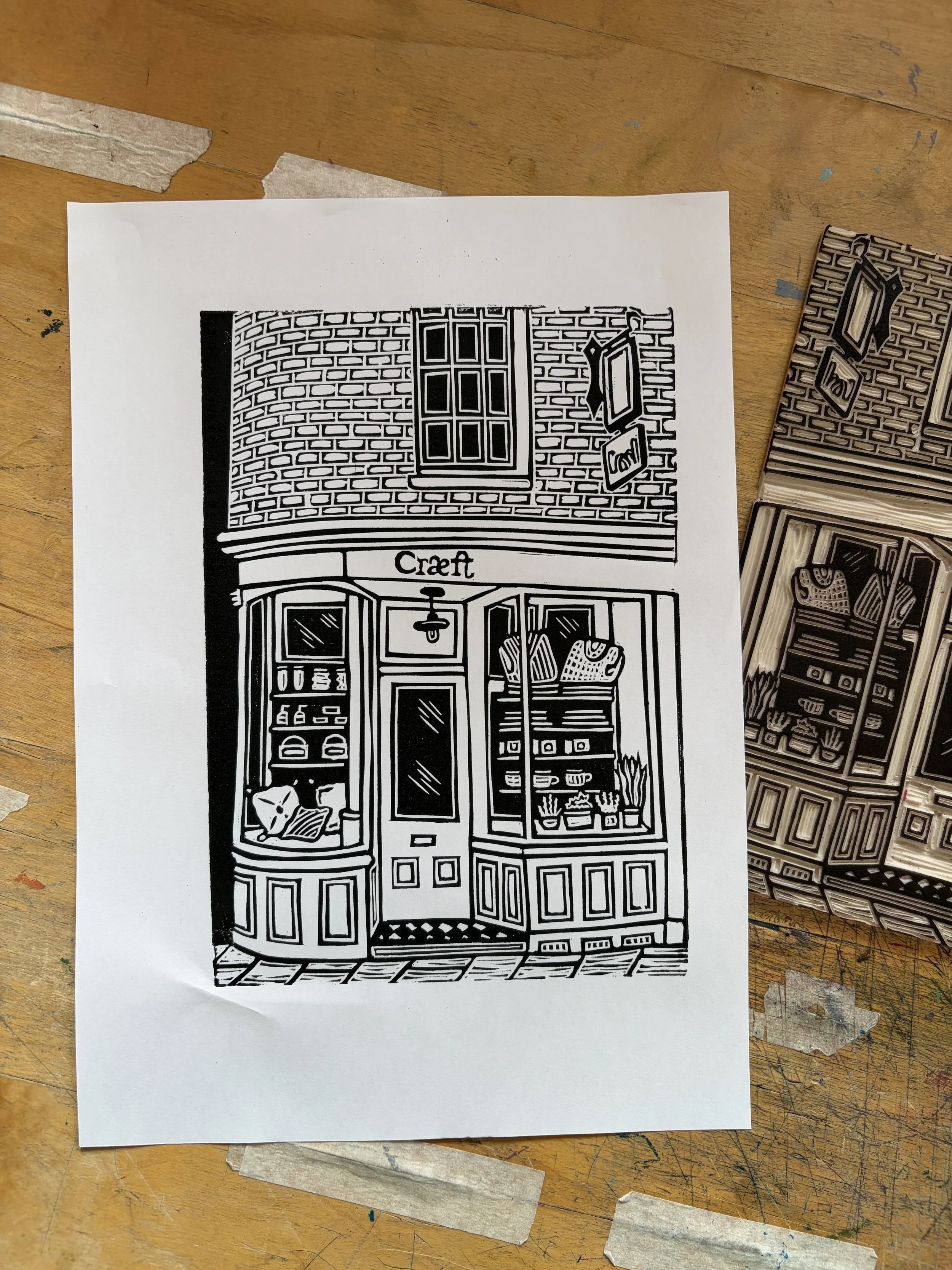 Black and white linocut print of a storefront with large windows, a door, and a sign that reads 'Craeft', with brick facade and interior decor visible through the windows.