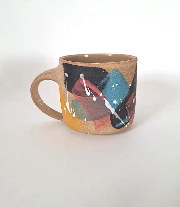 A ceramic mug with an abstract paint splatter design in red, black, blue, green, orange, and white on a neutral background.