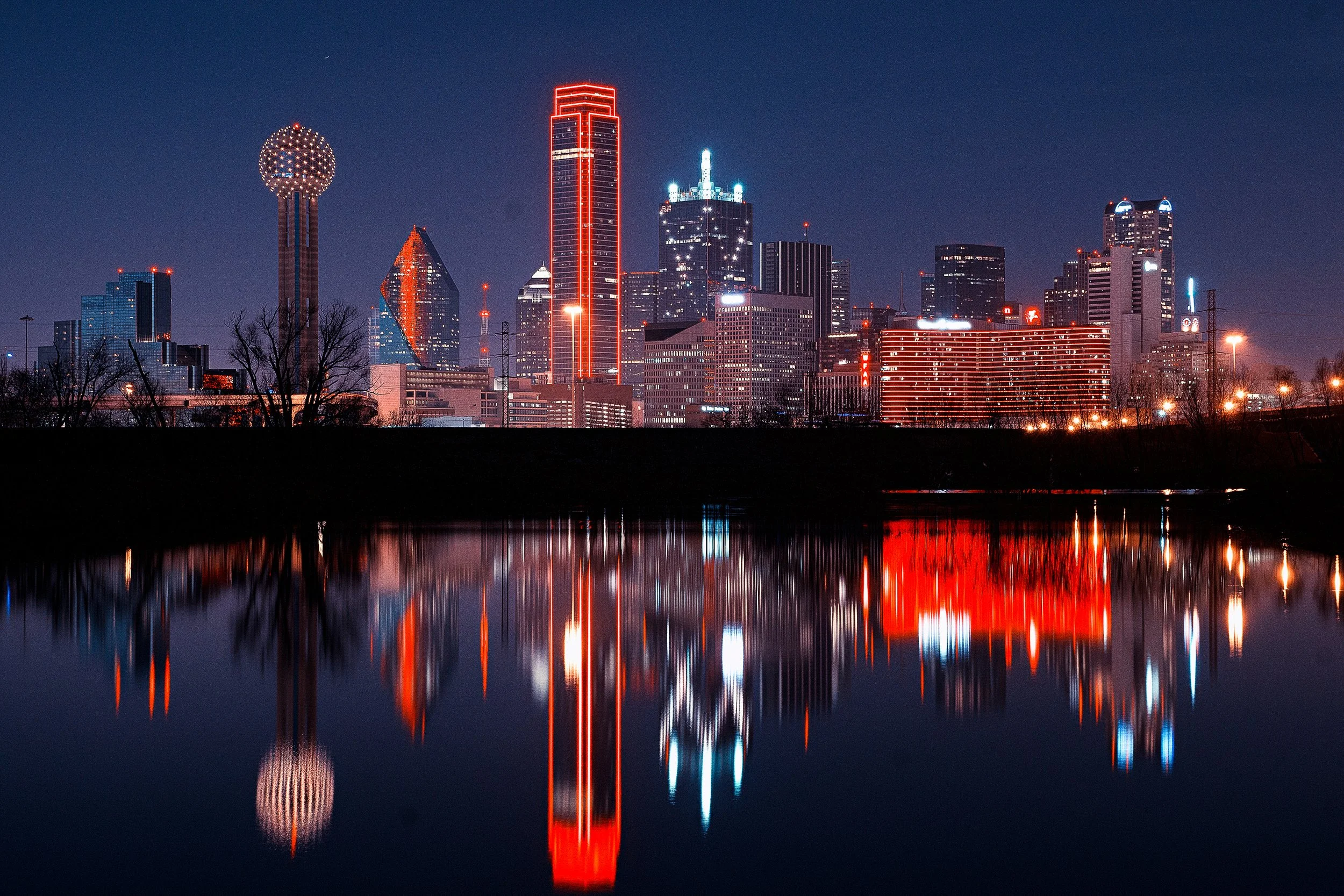 Night skyline of Dallas, Texas with illuminated skyscrapers reflected in a calm body of water.