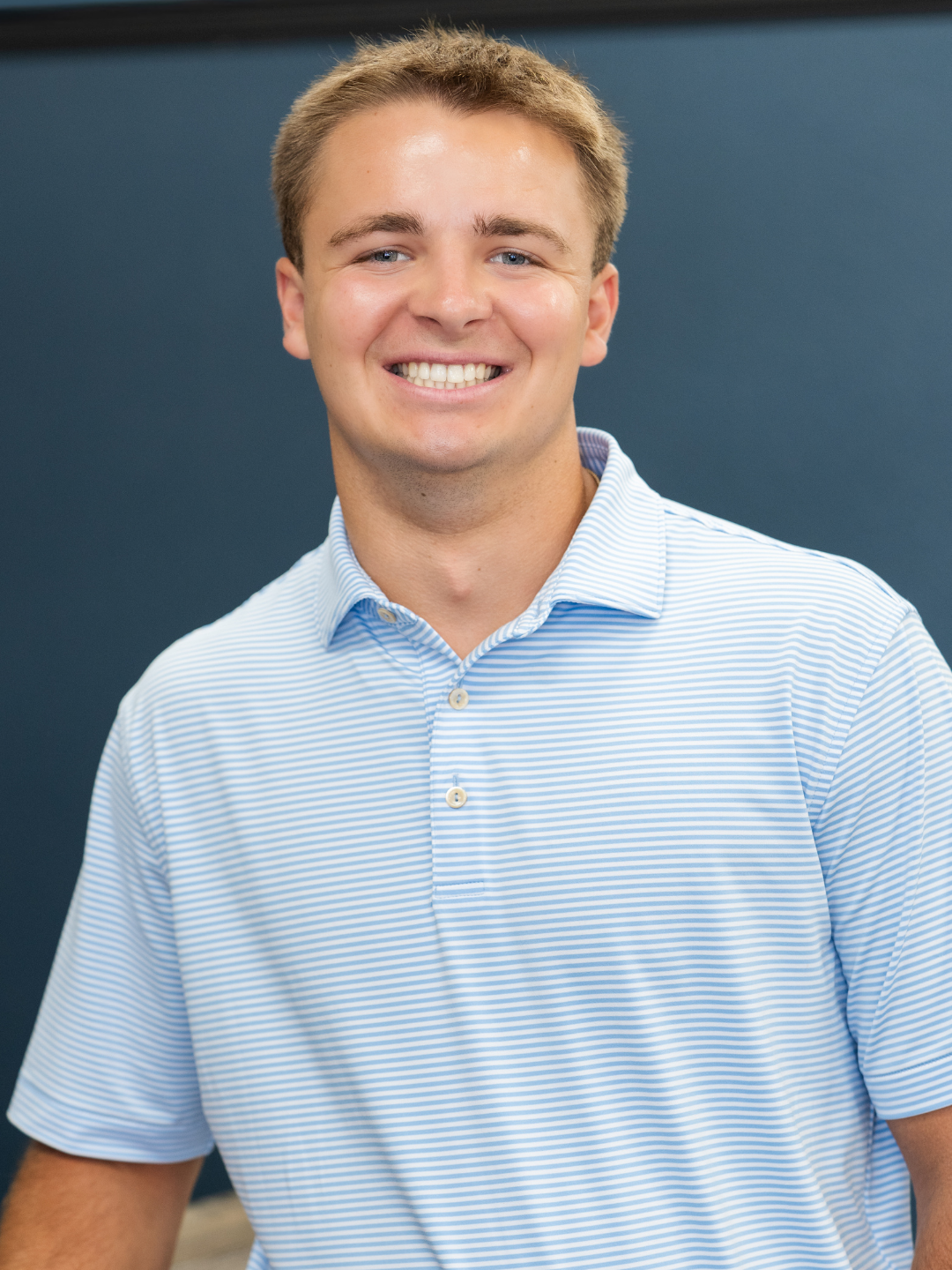 A young man with short, light brown hair, smiling, wearing a light blue and white striped collared shirt, standing against a dark blue background.