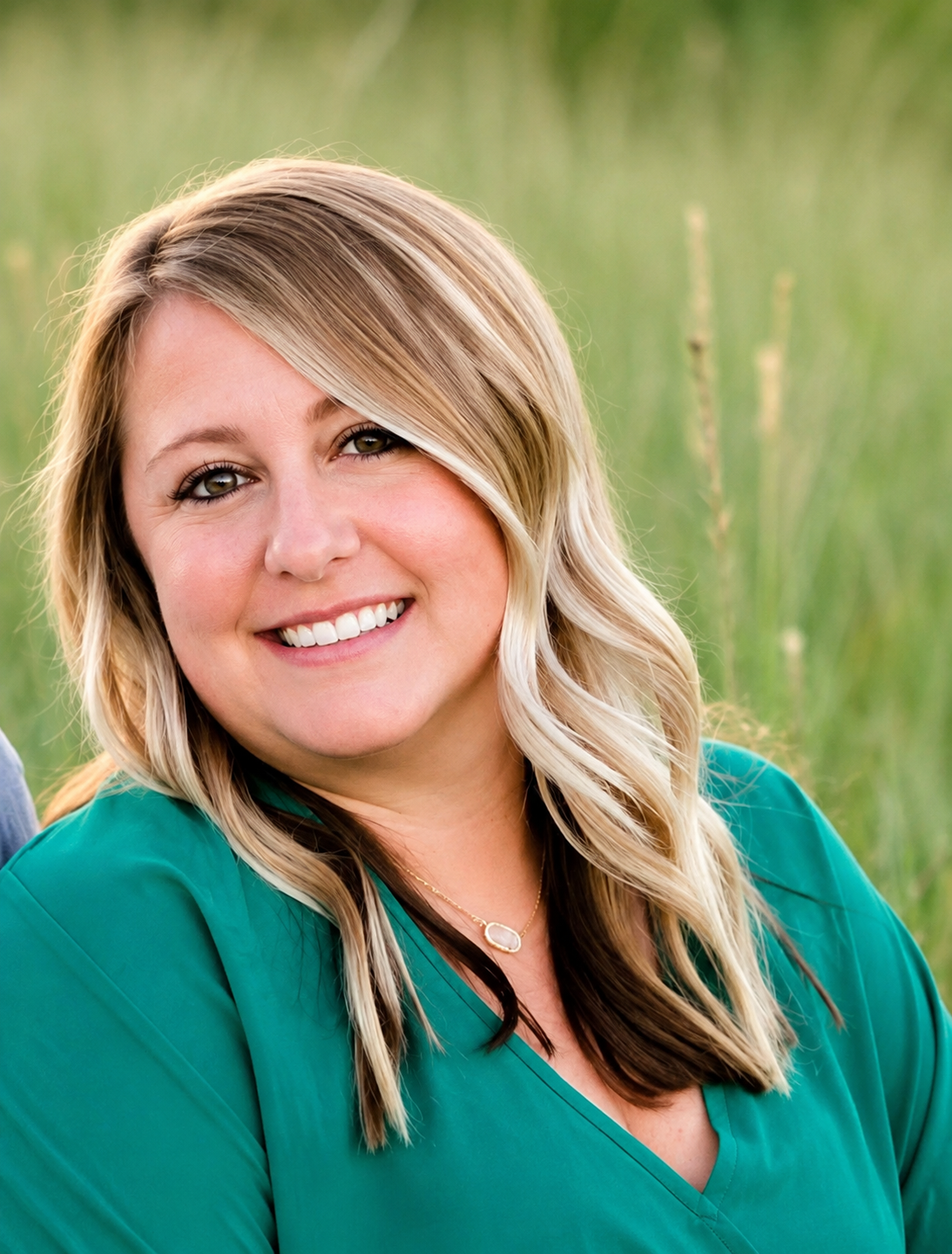 A woman with blonde, wavy hair smiling outdoors, wearing a green top and a gold necklace, with a blurred green field background.