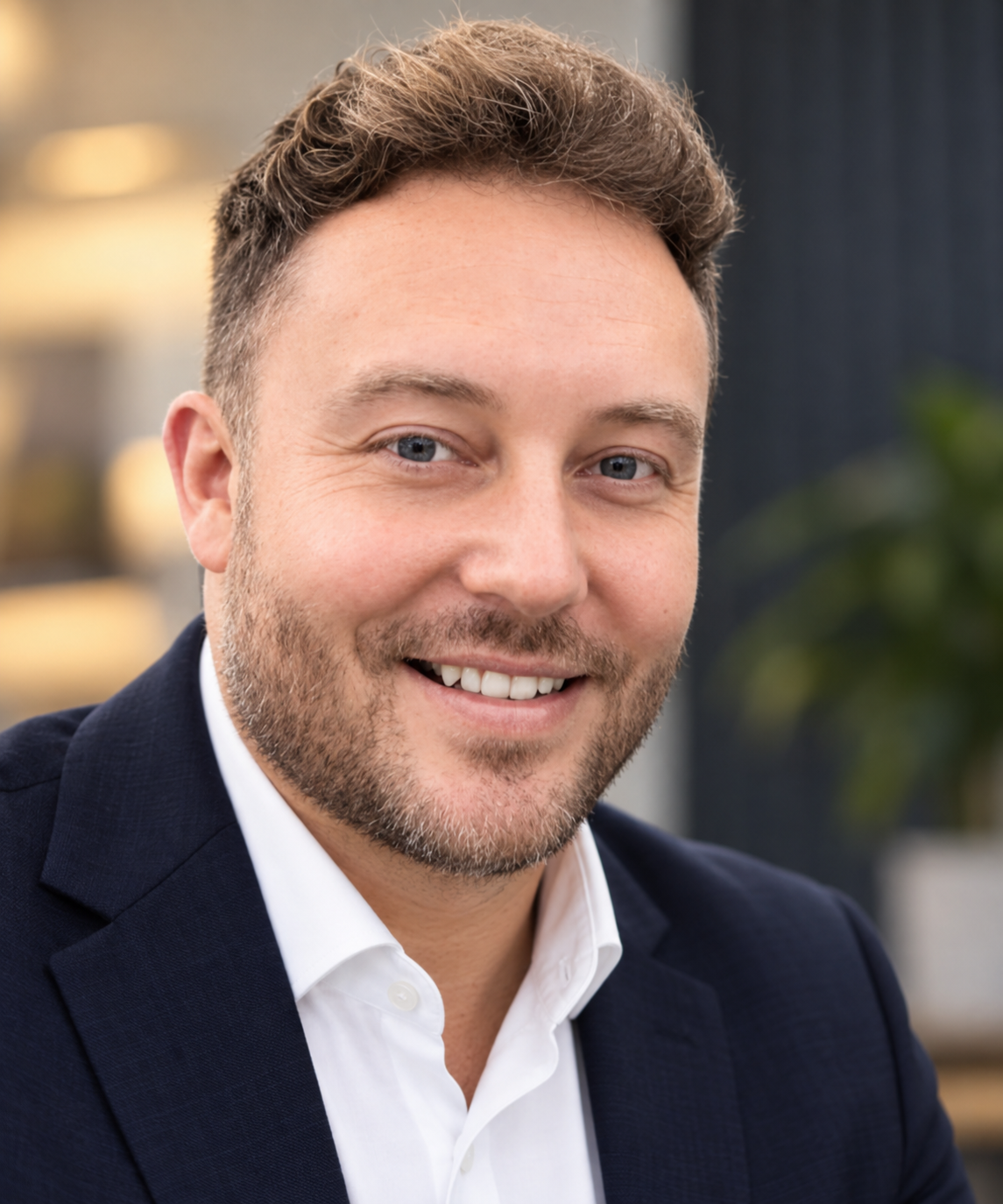 Close-up of smiling man Alistair West with light brown hair, beard, blue eyes, wearing a dark blazer and white shirt, in an indoor setting with blurred background.