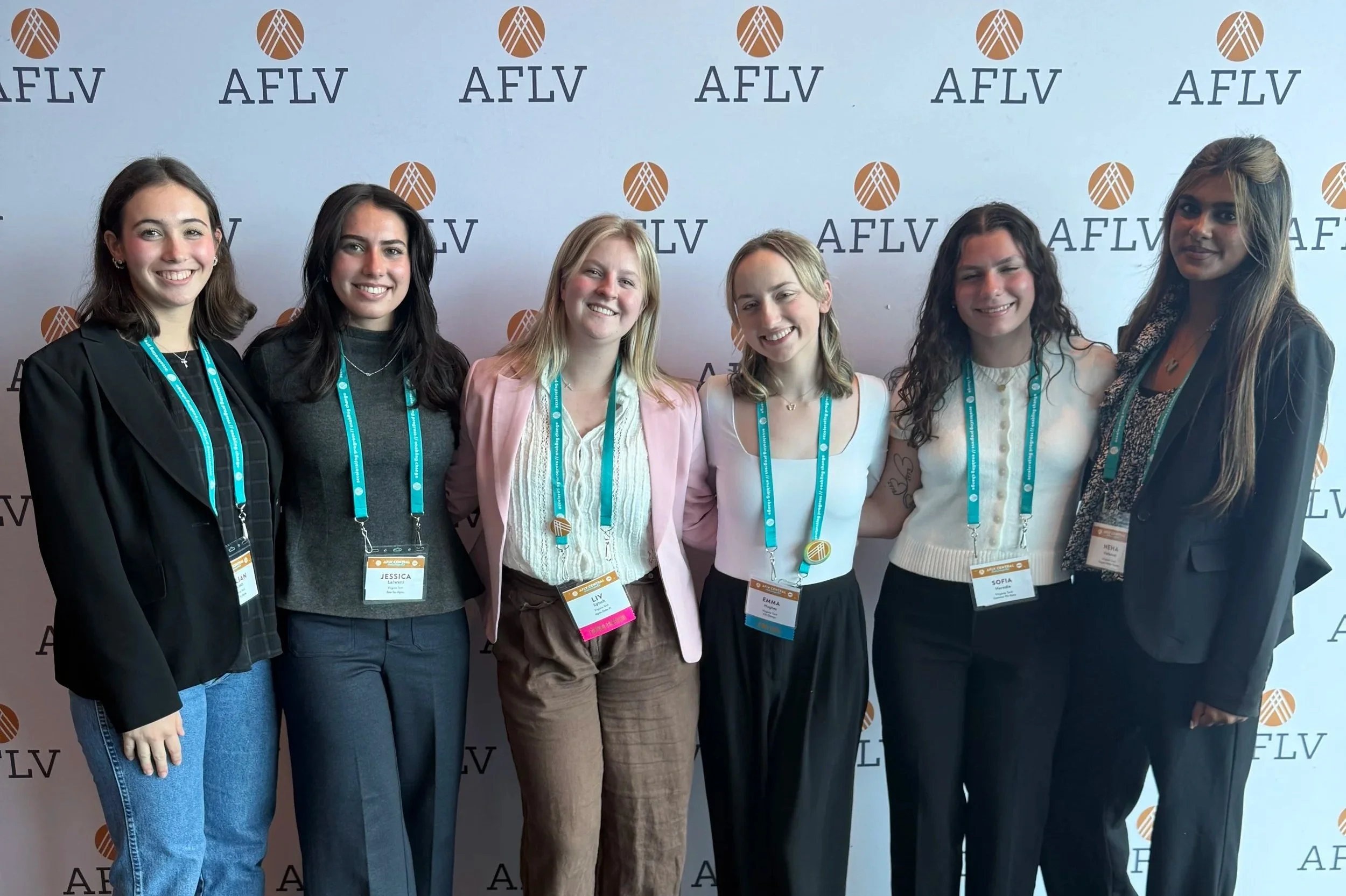 Group of six women standing together at an event, smiling, with a backdrop displaying the AFLV logo.