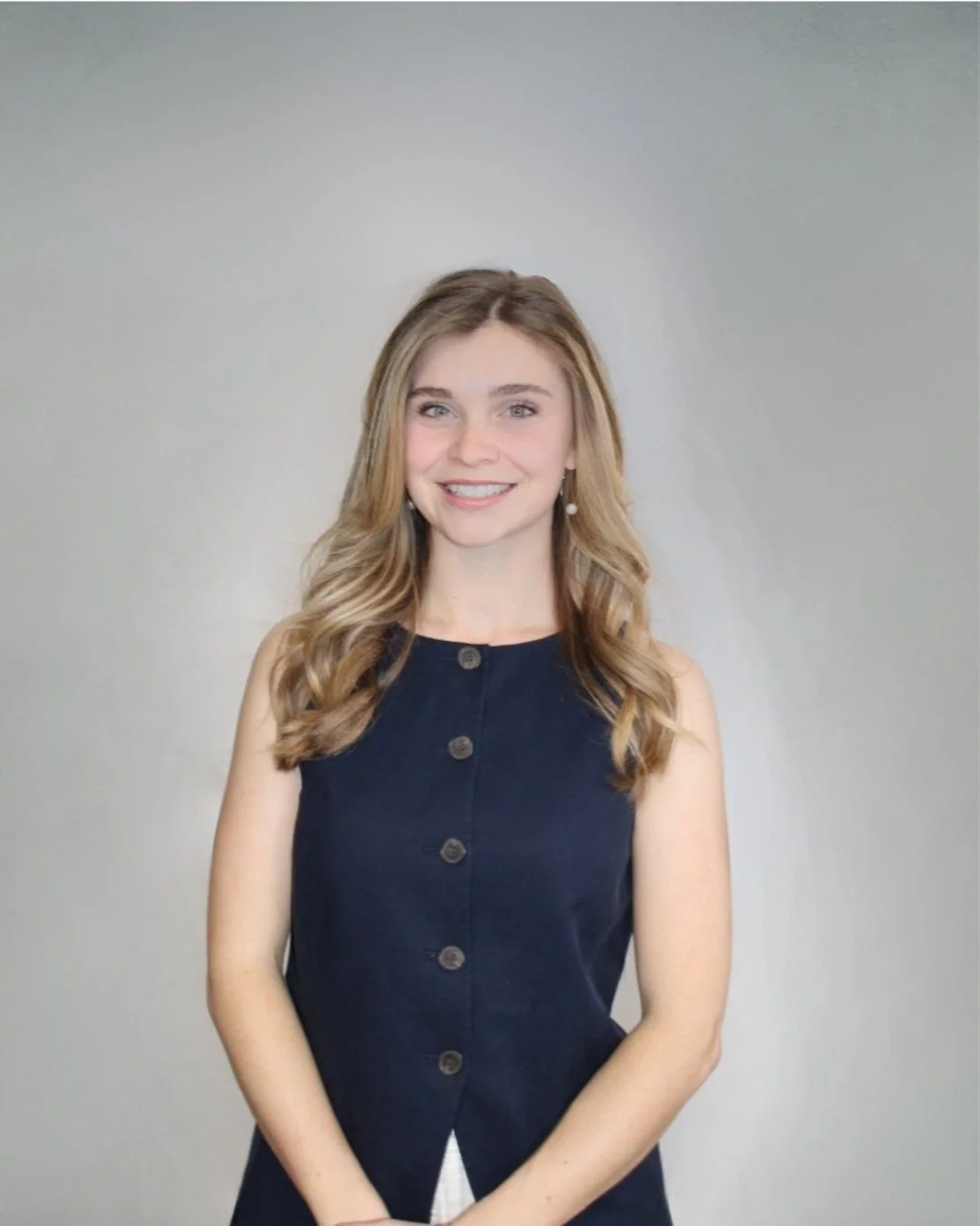 A young woman with long, wavy blonde hair smiling, wearing a sleeveless navy blue dress with large buttons, white earrings, standing against a plain, light gray background.