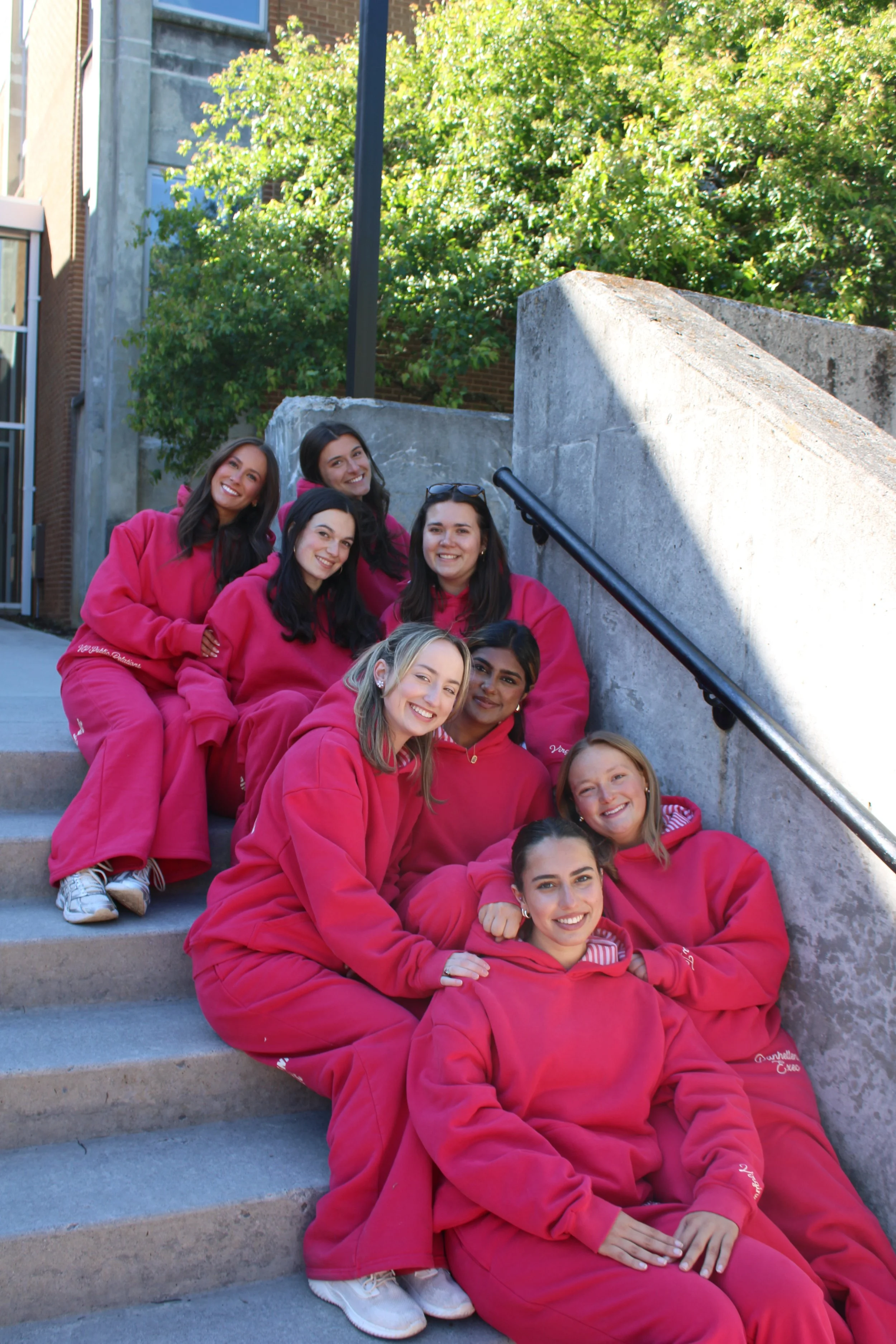 Group of young women wearing matching pink sweatshirts and sweatpants sitting on stairs outside, smiling for the camera.
