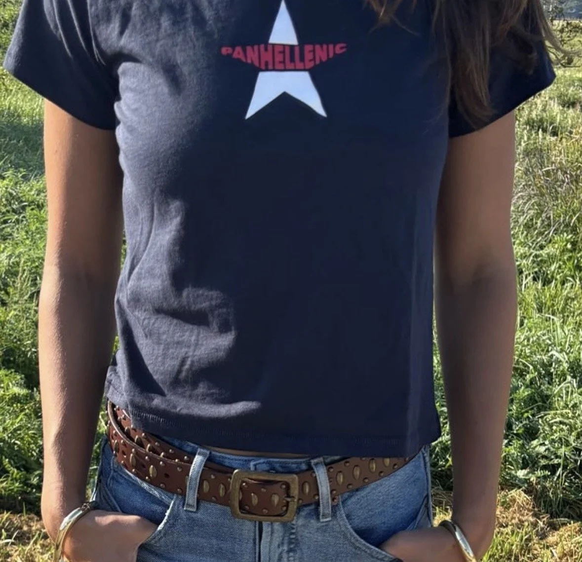 A person wearing a navy blue t-shirt with a white star and red text that reads 'PANHELLENIC' on the front. They are also wearing blue jeans, a brown studded belt, and standing outdoors in a green, grassy area.