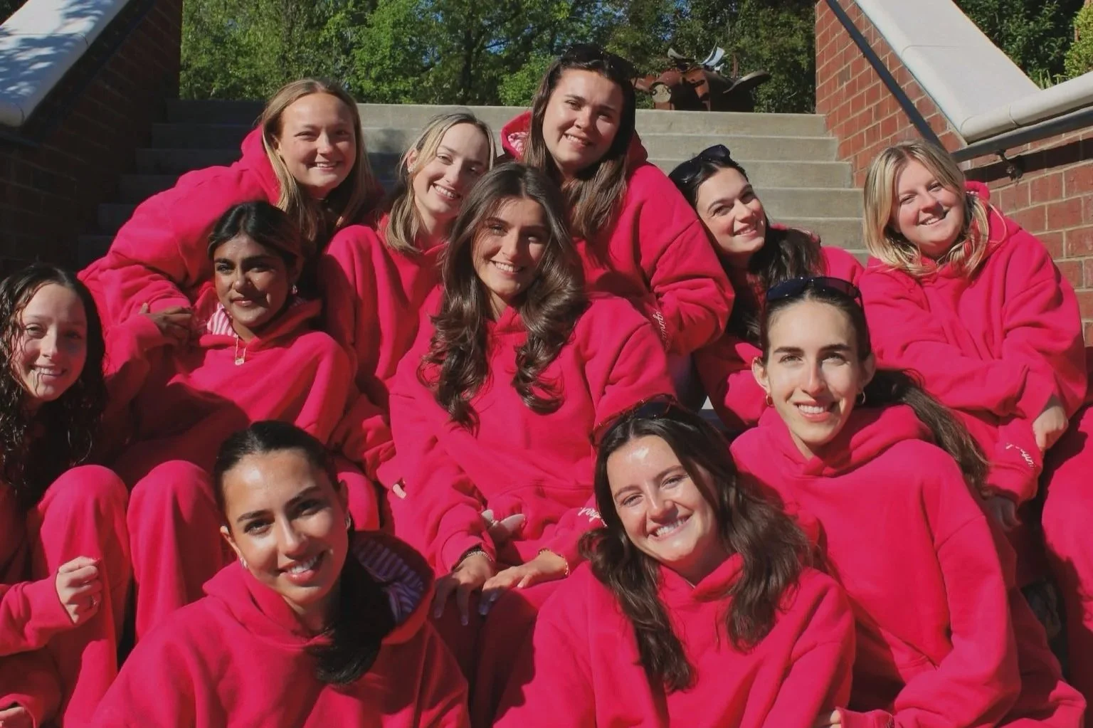 A group of women dressed in matching bright pink hoodies and pants sitting and standing on outdoor stairs, smiling for the camera.