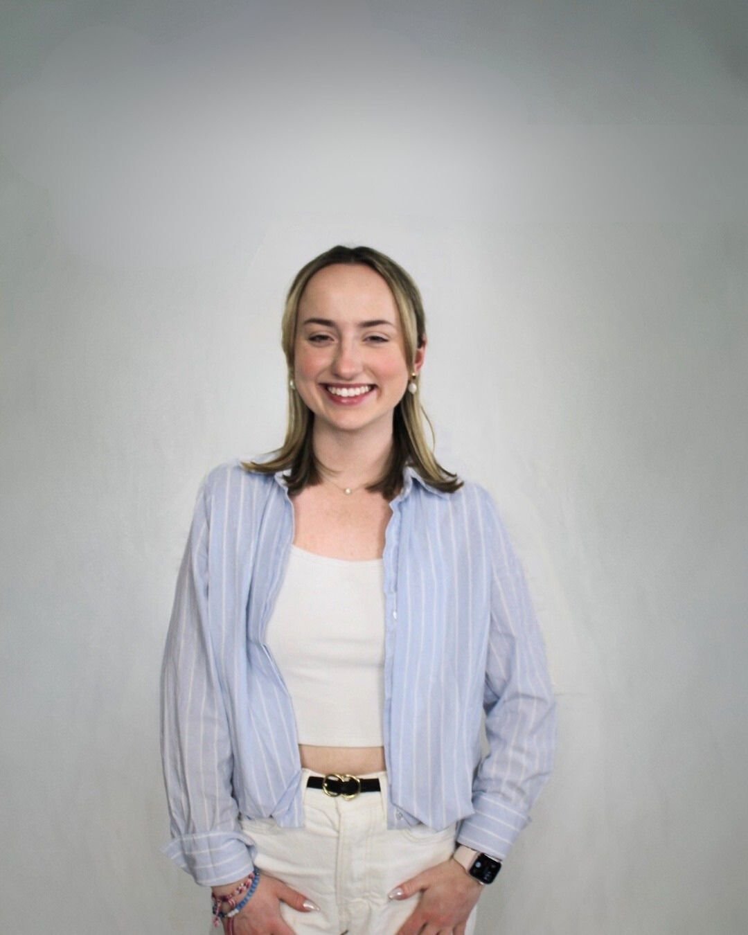 A young woman with shoulder-length blonde hair, smiling, wearing a light blue and white striped shirt over a white crop top, beige pants, and jewelry, standing against a plain light-colored background.
