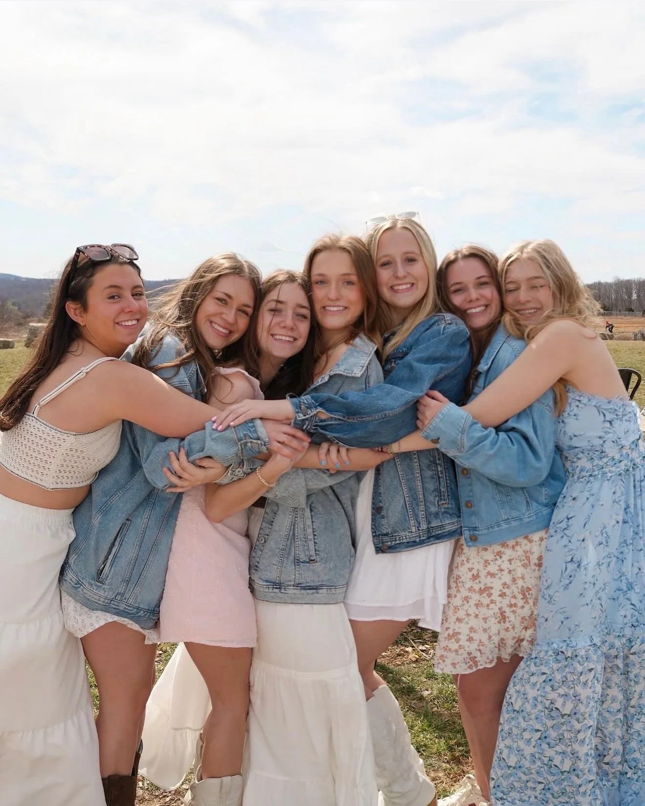 Group of seven young women hugging and smiling outdoors on a cloudy day, dressed in light summer clothing and denim jackets, with open field and trees in the background.