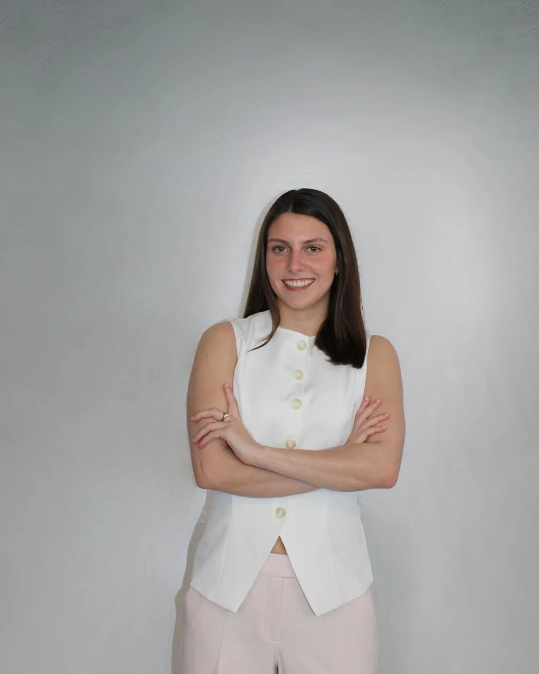 A young woman with brown hair in a white sleeveless top with buttons and light pink pants, smiling and crossing her arms, standing against a plain gray background.