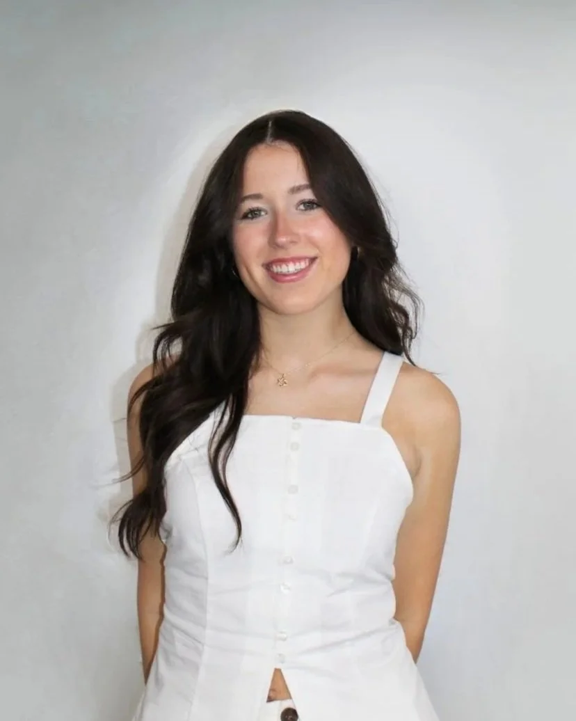 A young woman with long dark hair, smiling, wearing a white sleeveless button-up dress against a plain white wall.