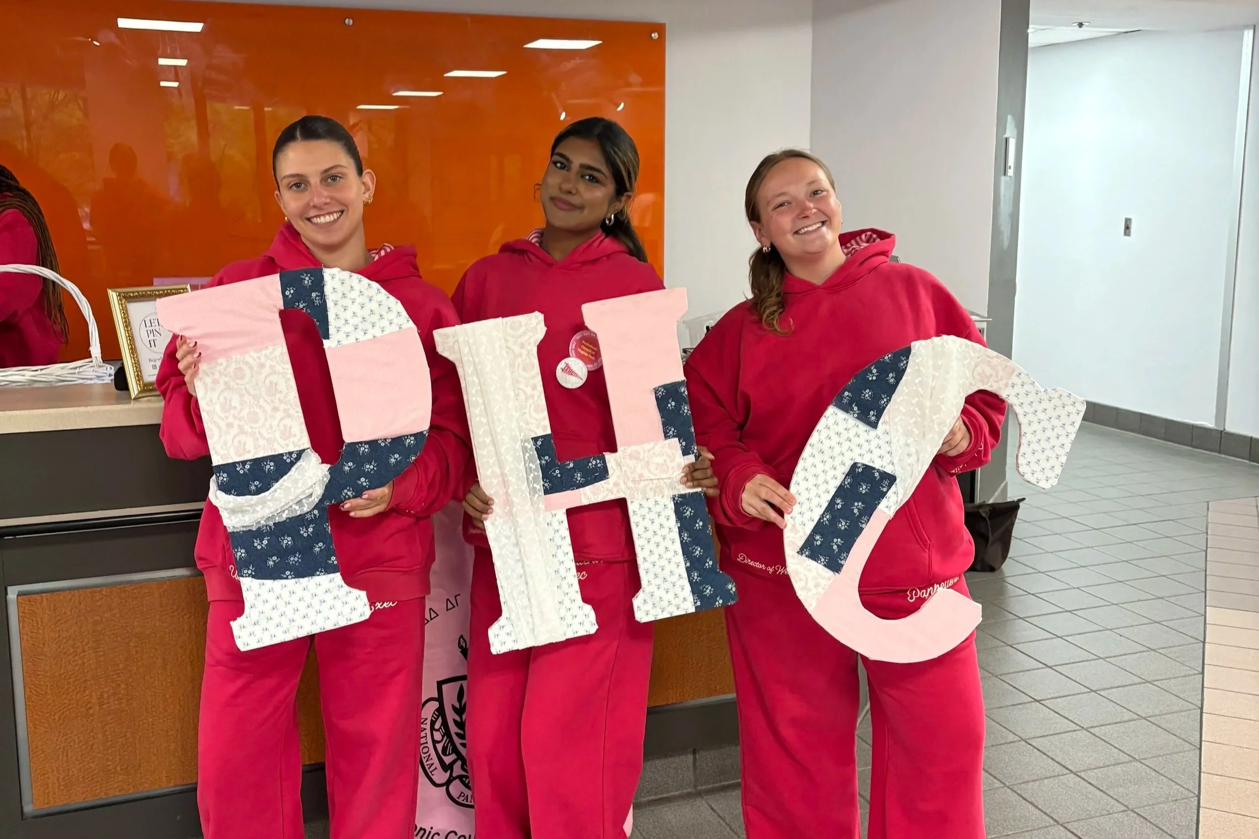 Three women standing indoors, smiling, holding large letters spelling 'LIFT' decorated with pink, white, and blue patterned fabric, wearing matching red hoodies and pants.