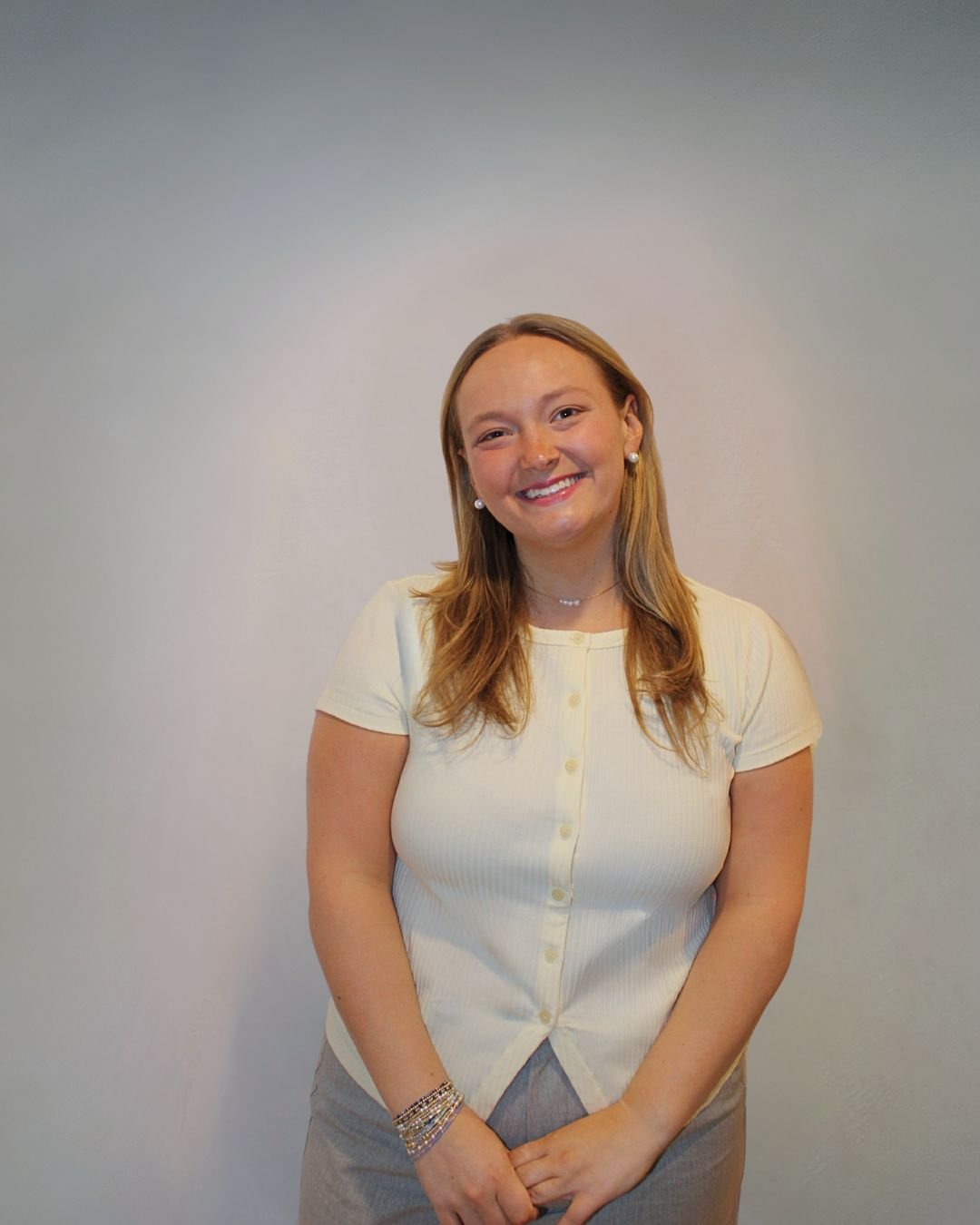 Smiling woman with shoulder-length hair, wearing a light yellow button-up shirt, jewelry, and standing in front of a plain wall.