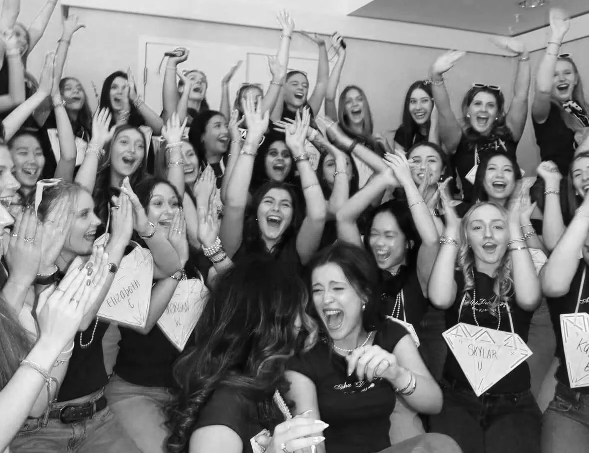 A group of young women celebrating and laughing together at a party or event, some wearing name tags and jewelry, in a joyful and lively atmosphere.