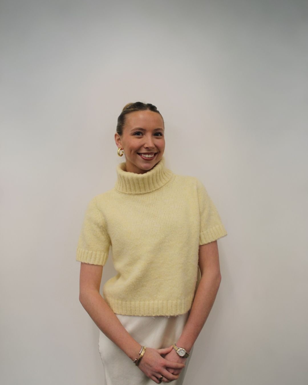 A woman with light skin and blonde hair tied back, wearing gold hoop earrings, a gold bracelet, and a watch, stands against a plain white wall. She is smiling and wearing a yellow turtleneck sweater with short sleeves, and light-colored pants.