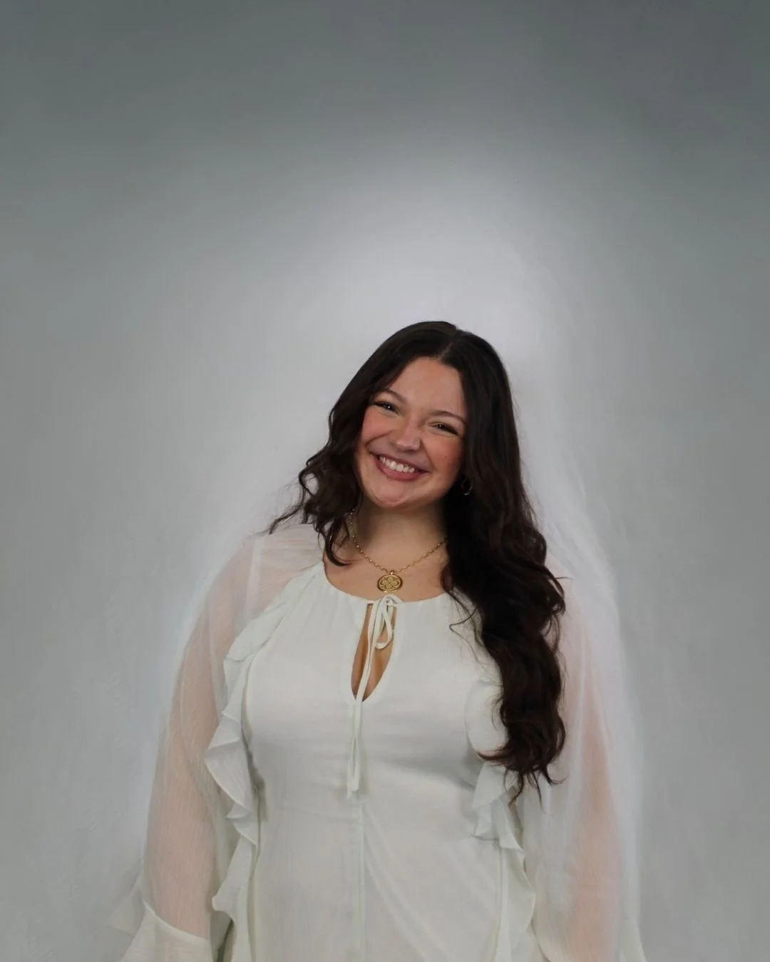 A young woman with long dark wavy hair, smiling, wearing a white blouse with ruffles and a tie at the neck, and a gold necklace with a pendant, standing against a plain gray wall.