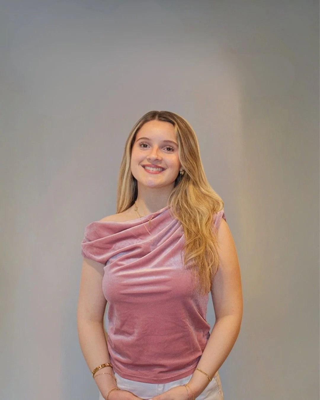 A young woman with long, wavy blonde hair smiling while standing against a plain gray background, wearing a pink velvety off-shoulder top and white pants.