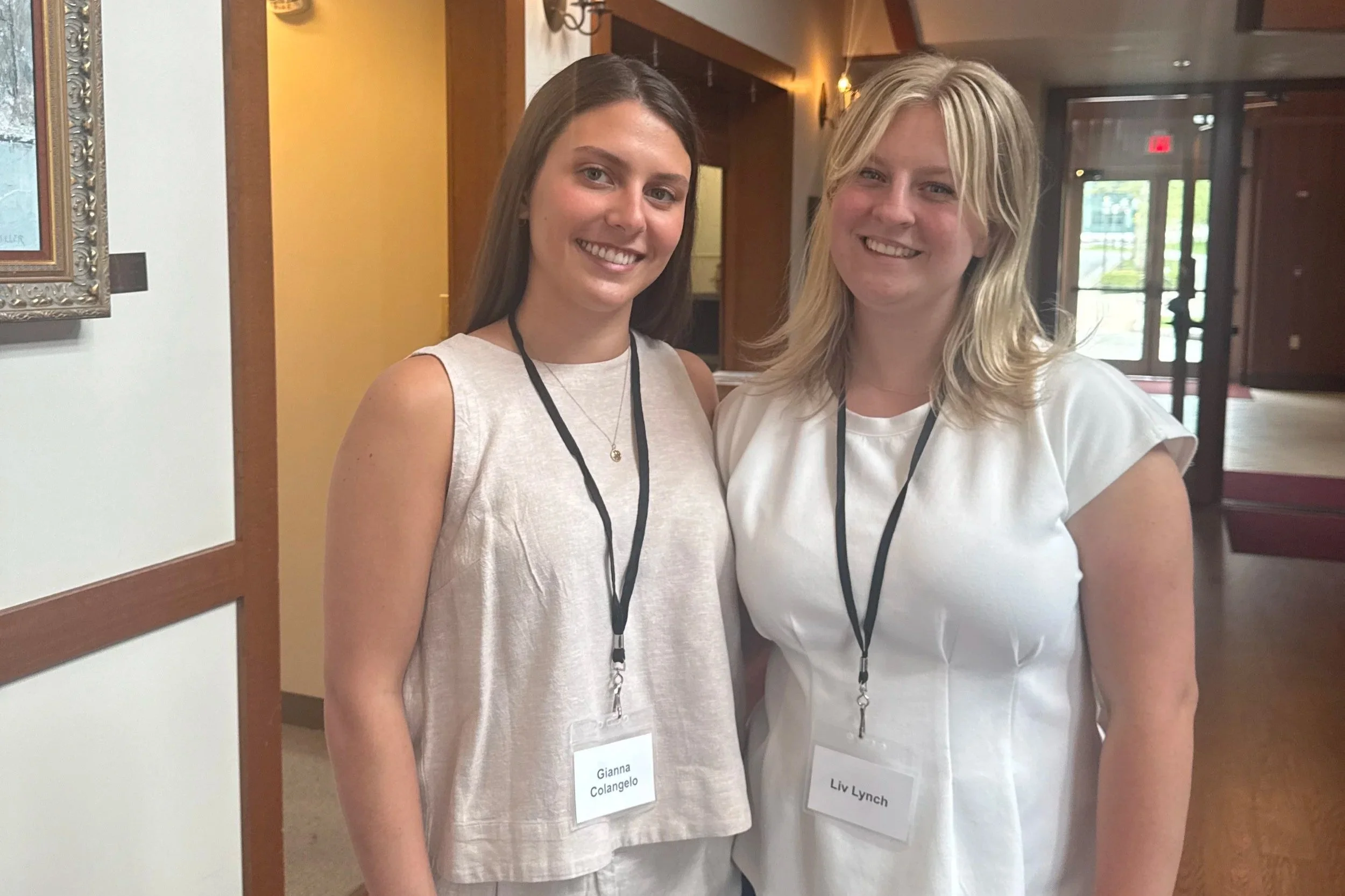 Two women standing side by side indoors, both wearing white tops and conference name tags on black lanyards. The woman on the left has brown hair, and the woman on the right has blonde hair. They are smiling at the camera.