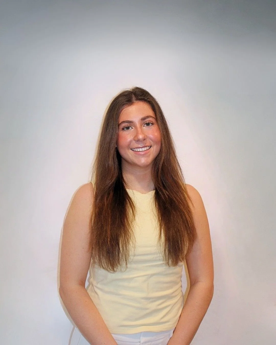 Young woman with long brown hair smiling, wearing a sleeveless yellow top, standing against a plain light-colored background.