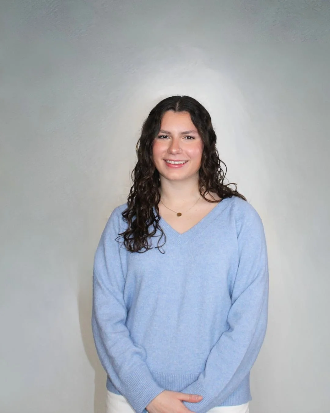 Young woman with long curly dark hair smiling, wearing a light blue V-neck sweater and a small gold pendant necklace, standing against a plain light gray background.