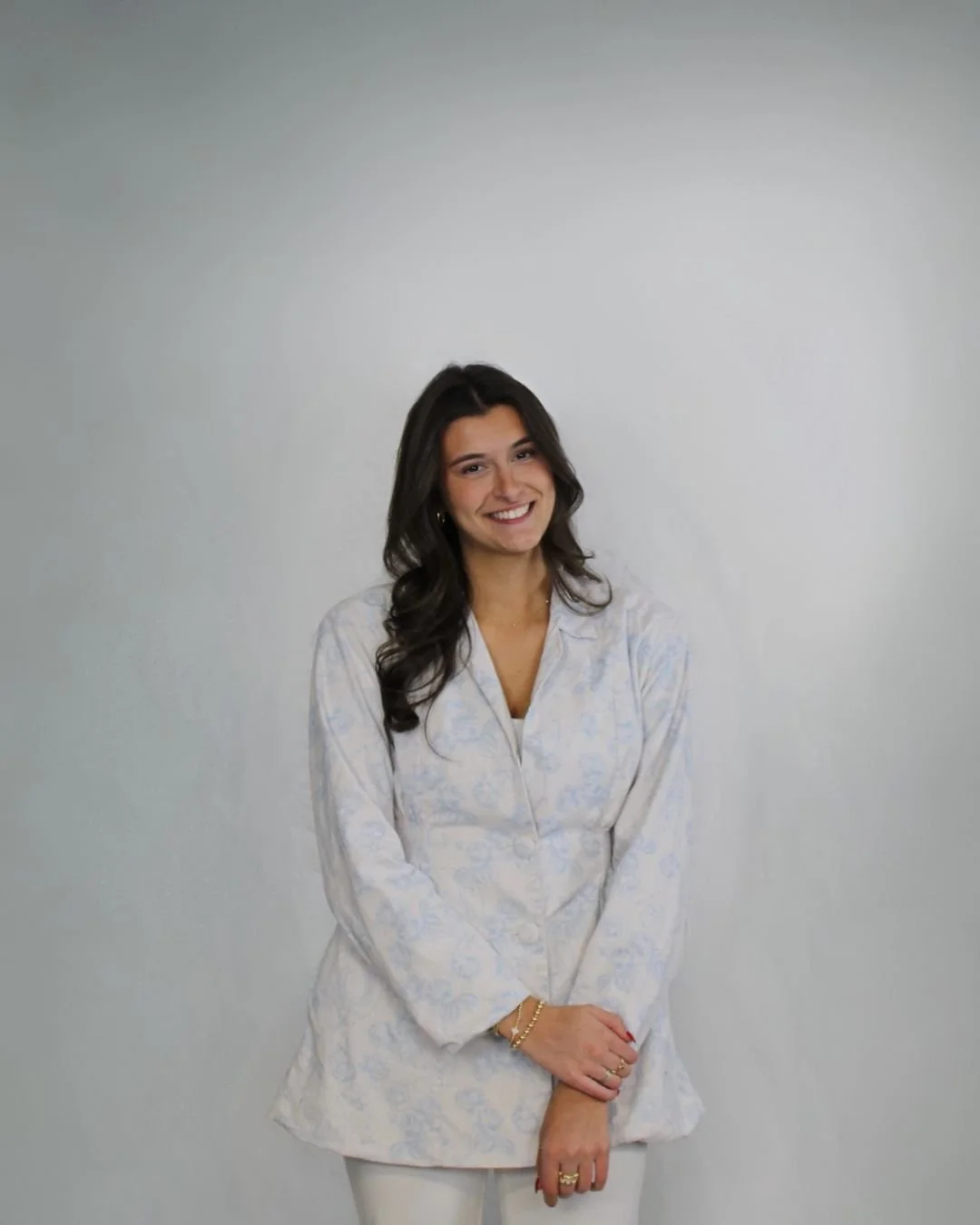 A young woman with long dark hair and a bright smile, wearing a light-colored patterned blouse and white pants, standing against a plain light gray wall.