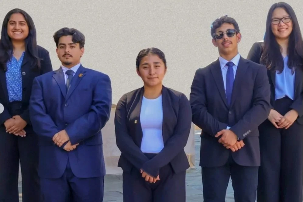 Group of five young adults, three women and two men, dressed in formal business attire, standing outdoors against a light-colored wall, smiling and posing for a photo.
