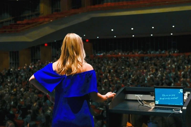 A woman with red hair wearing a blue dress stands on stage at a conference or presentation, facing a large audience seated in an auditorium.