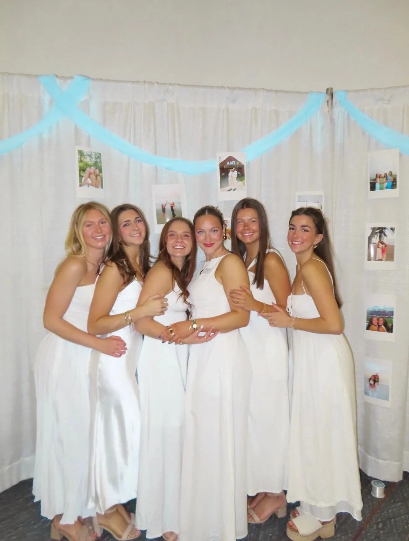 Six women in white dresses standing together at a celebration, posing for a photo with a backdrop of photographs and blue garland.