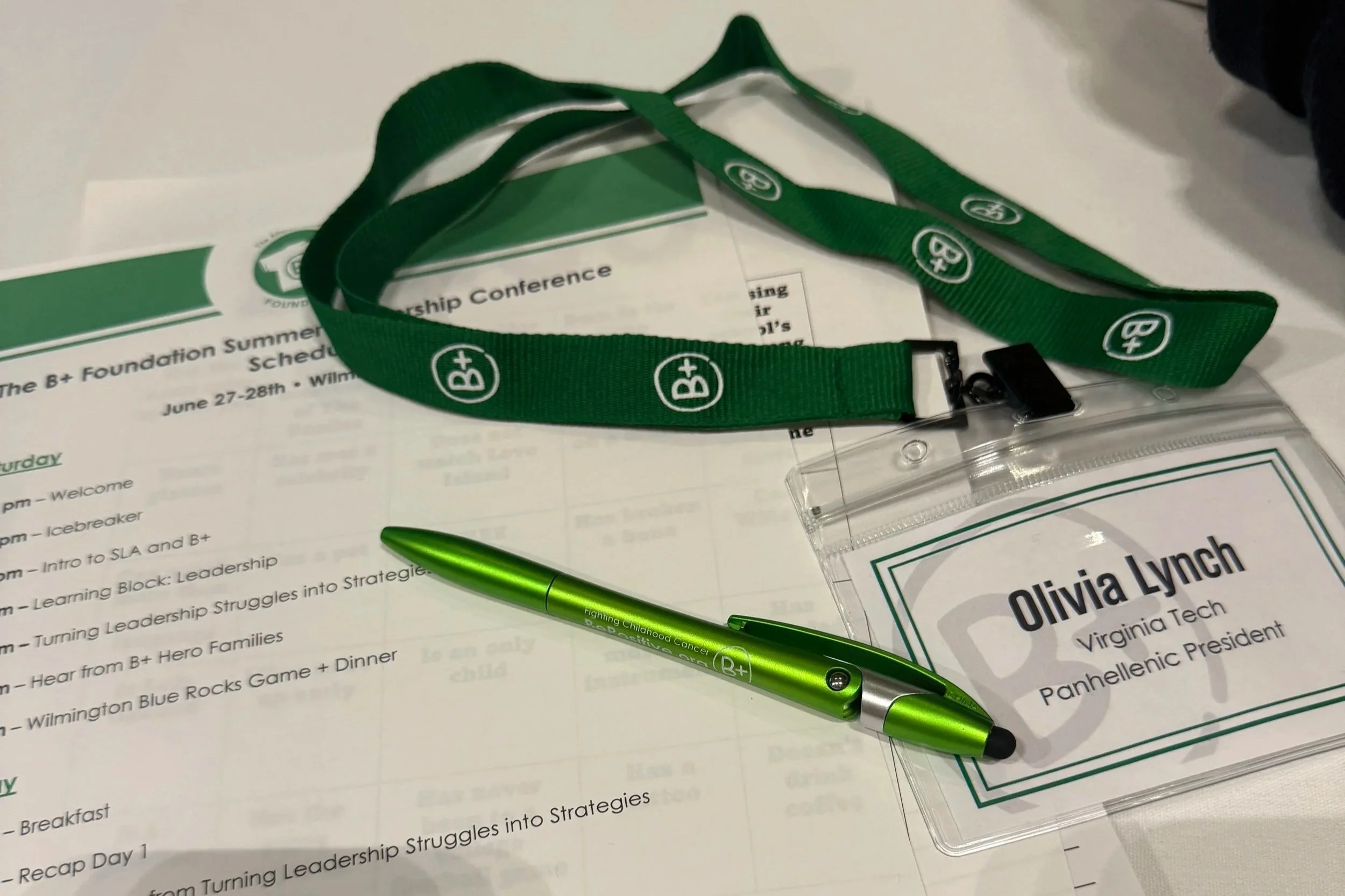 Conference materials including a green lanyard with B+ logo, a green and silver pen, and a name tag for Olivia Lynch at a leadership event.