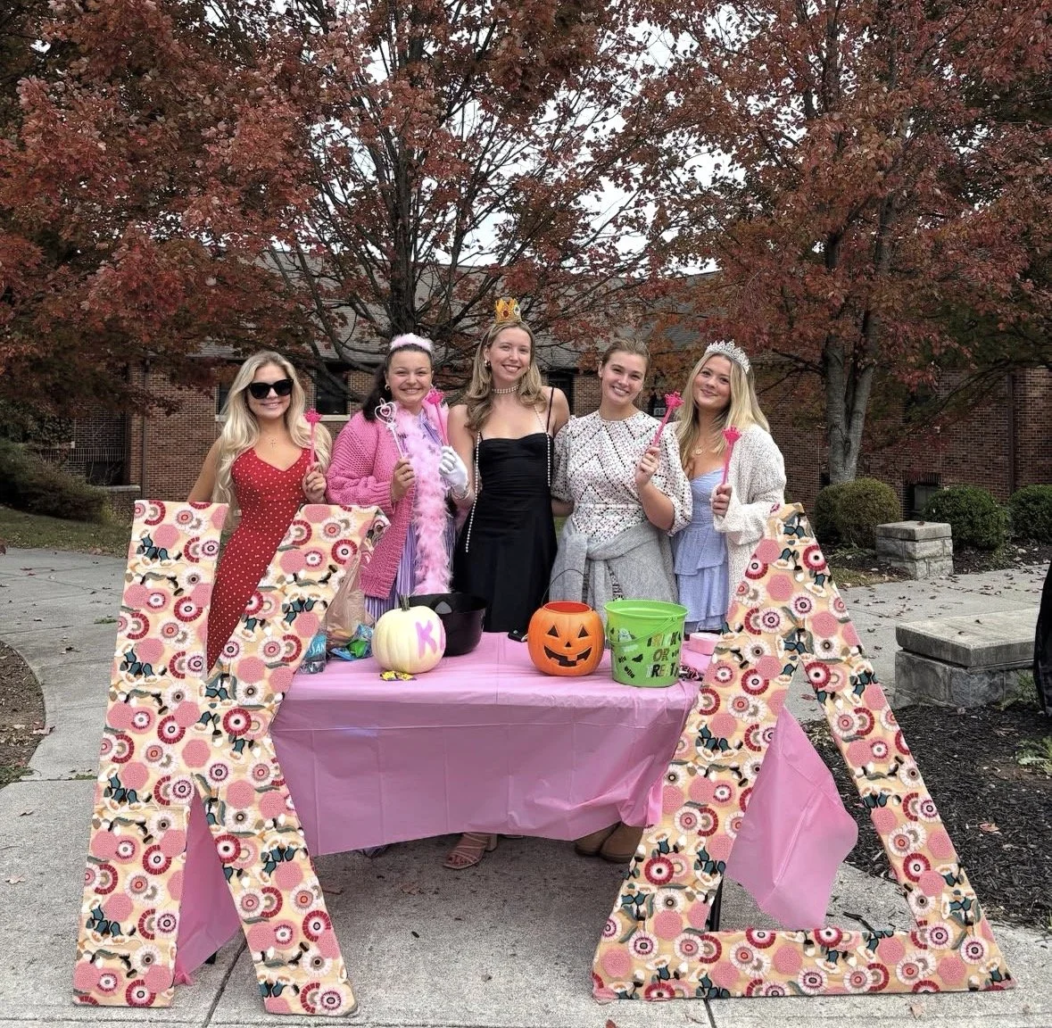 Five women dressed in colorful costumes and accessories, standing around a pink table with Halloween decorations, pumpkins, and buckets, outdoors during fall with trees showing red leaves.
