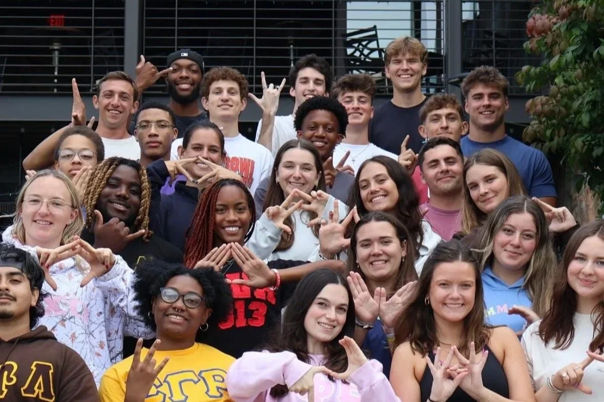 Group of diverse young people smiling and posing together outdoors, some making hand gestures.