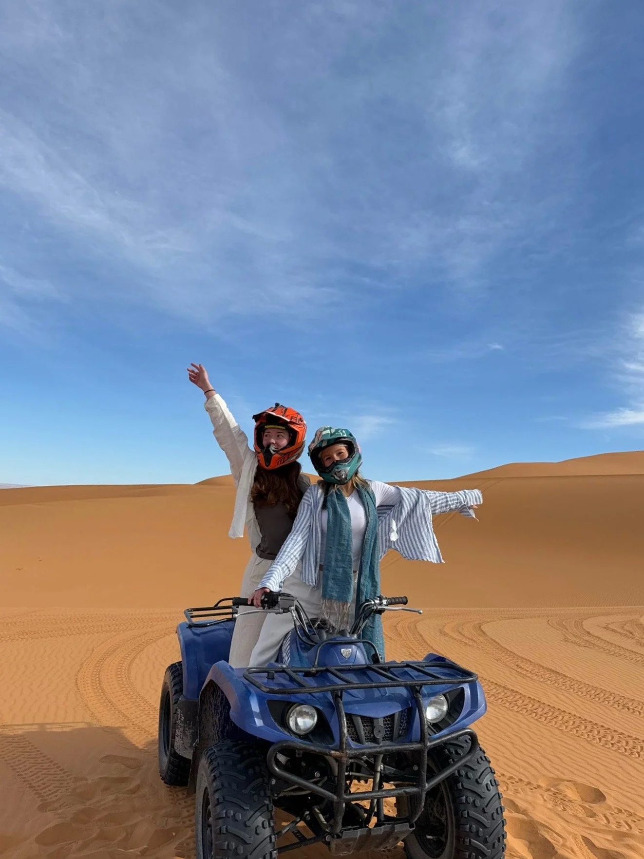 Two women wearing helmets and scarves riding a four-wheeler in a desert with sand dunes and a blue sky