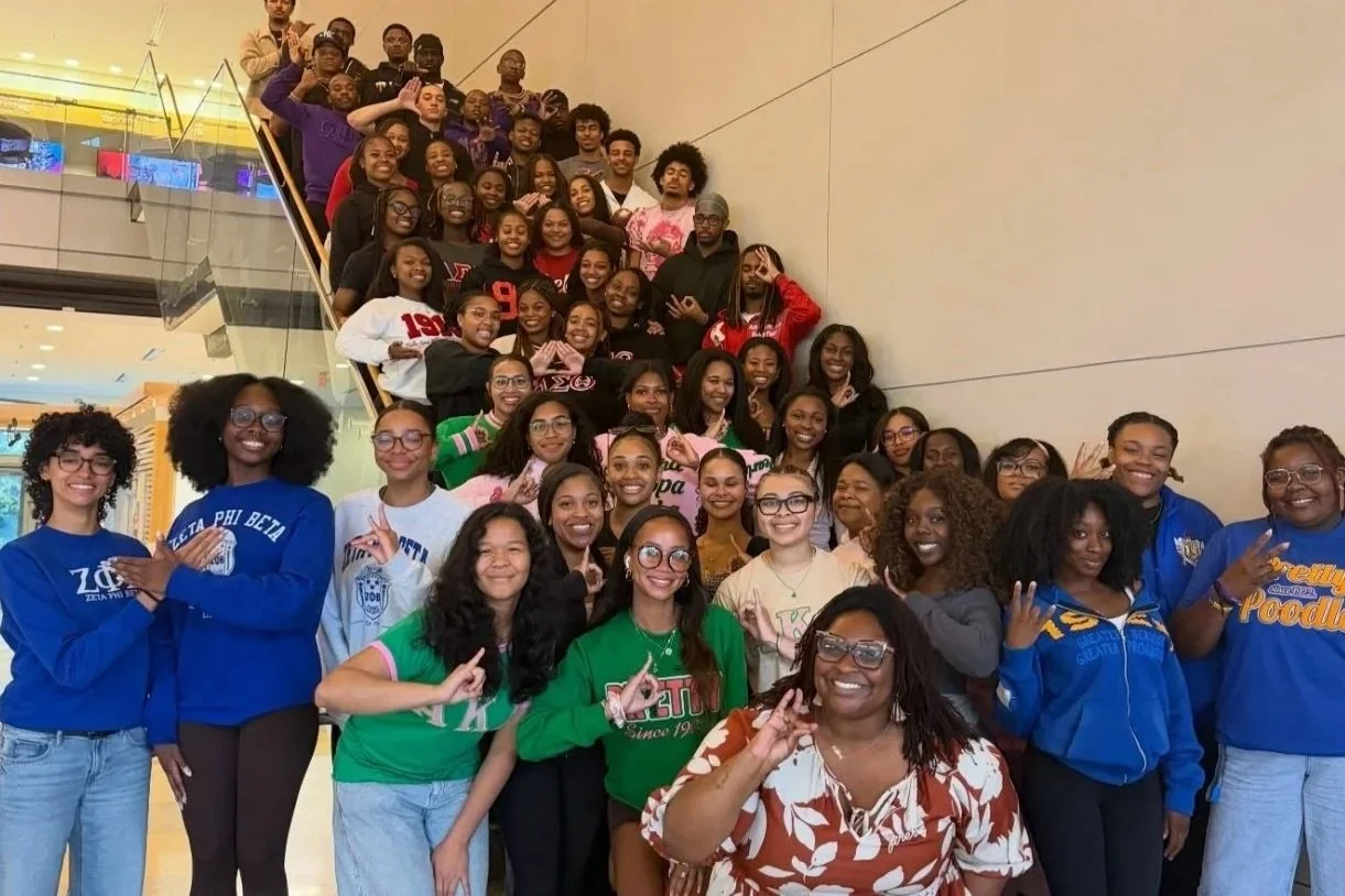 A large group of young women and a few men posing on a staircase inside a shopping mall or similar venue. Many are smiling and making hand gestures, and some are wearing fraternity or sorority shirts.