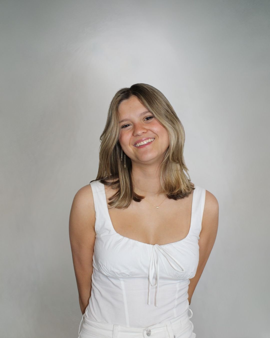 A young woman with shoulder-length blonde hair smiling, wearing a white sleeveless top and earrings, standing against a plain gray background.