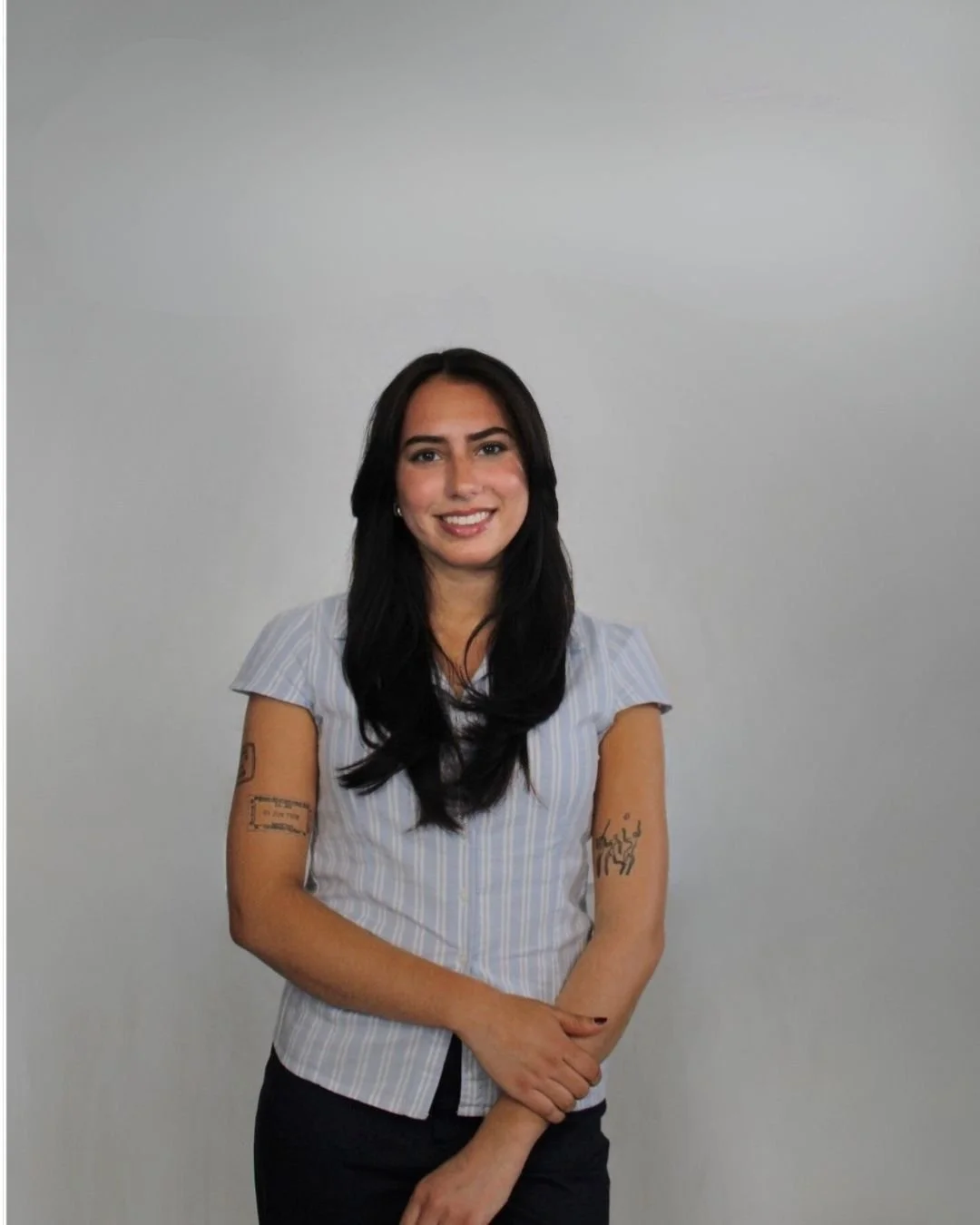 A smiling woman with long black hair, wearing a light blue striped shirt and black pants, standing against a plain light gray background.