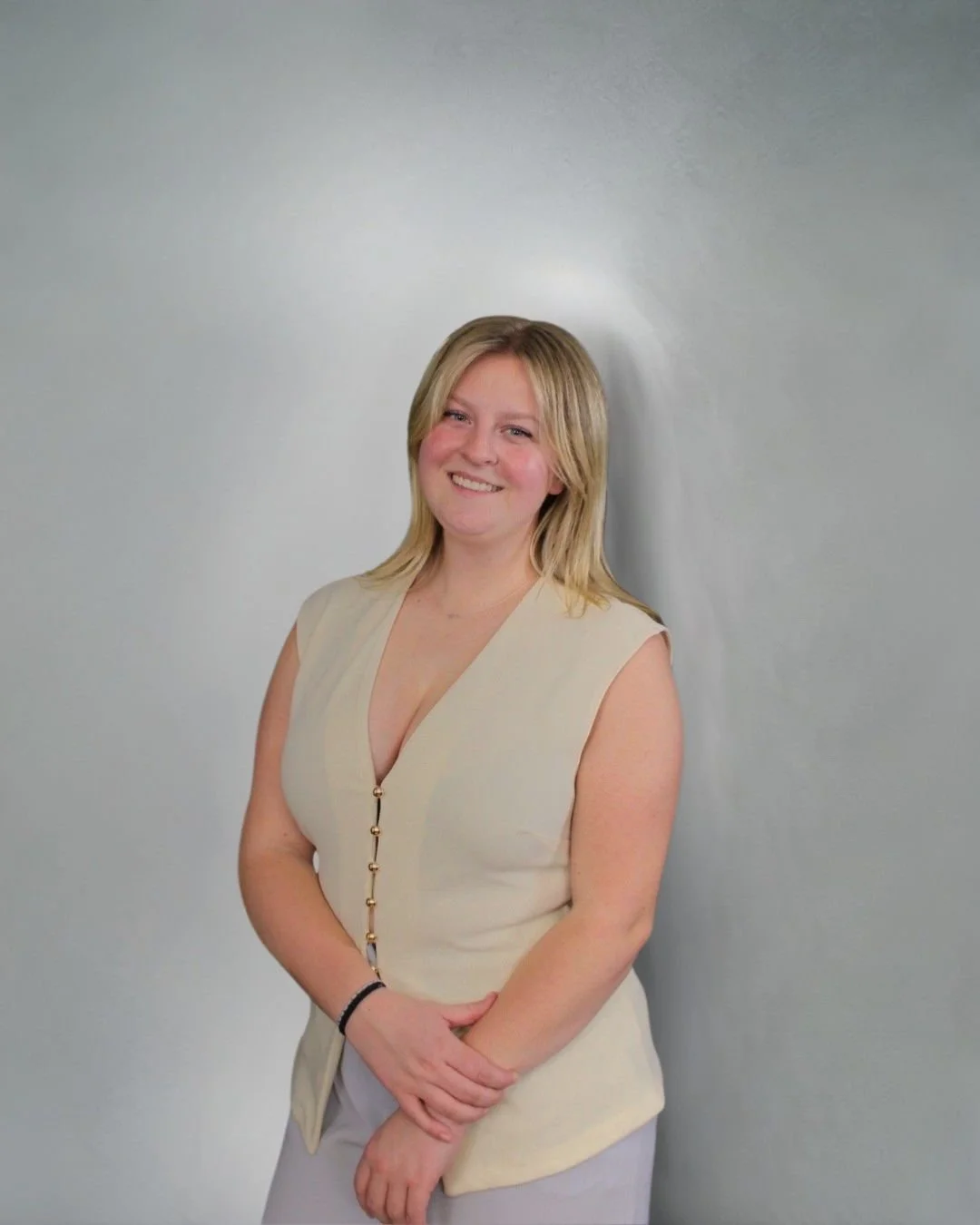 A young woman with blonde hair smiles while standing against a plain light gray wall, wearing a beige sleeveless top with a zipper down the front and light-colored pants.