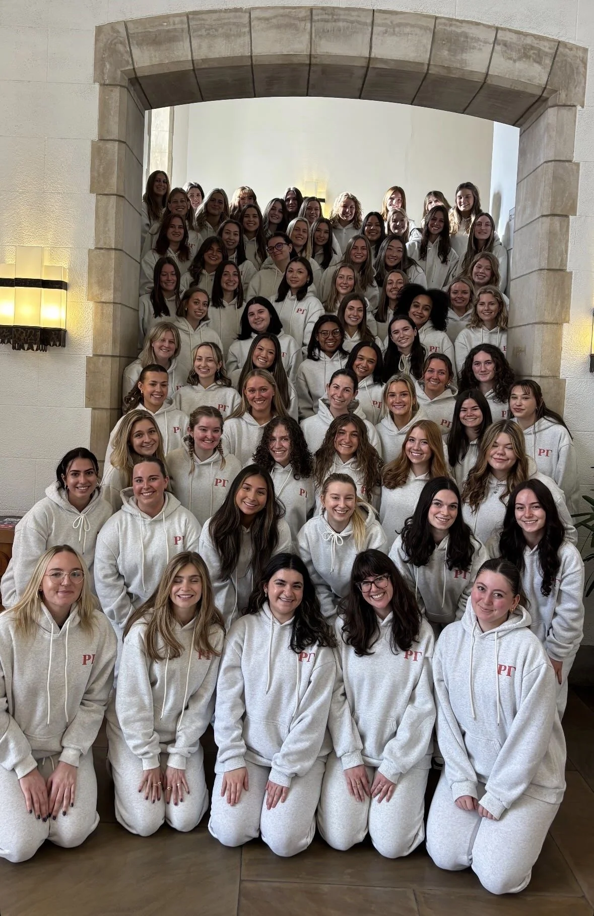 Group of young women wearing matching white hoodies with red initials, posing together indoors in front of a stone archway.
