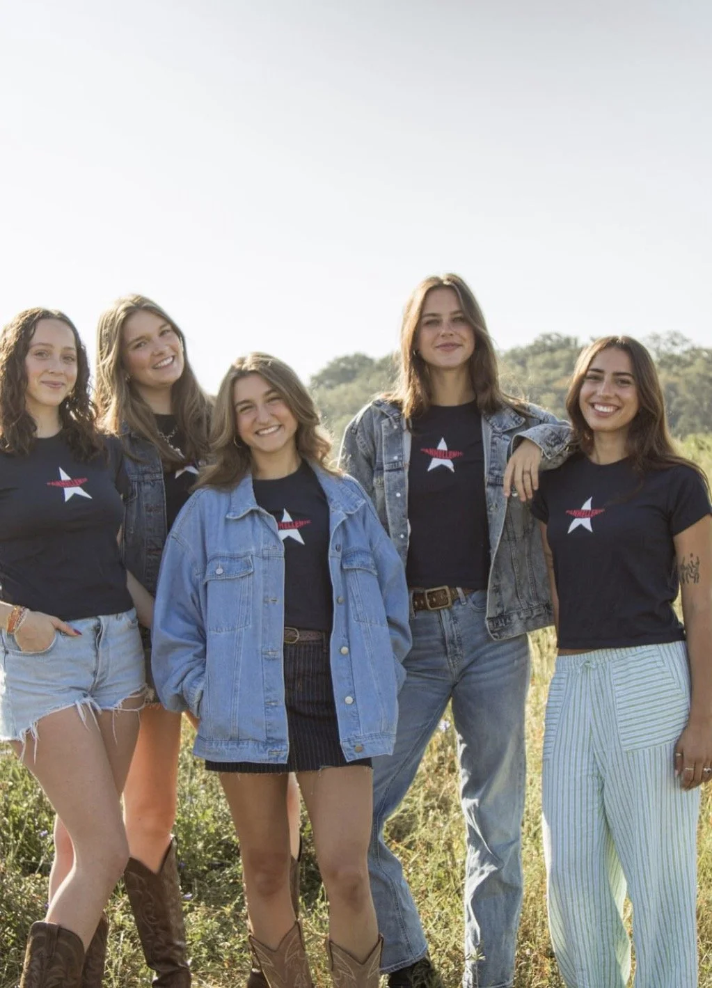 Group of five young women outdoors during daytime, wearing casual clothing with some in matching T-shirts with a star and red banner design, standing on grass with a backdrop of trees and a clear sky.