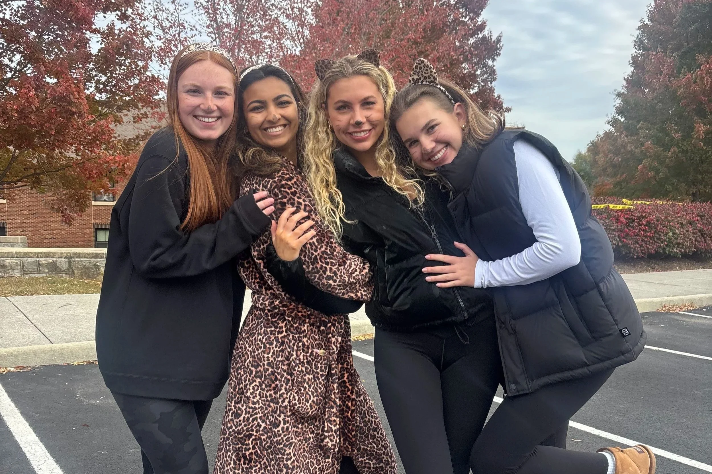 Four young women dressed in animal print and black jackets, wearing leopard ears and smiling, standing closely together outdoors with autumn trees and a brick building in the background.
