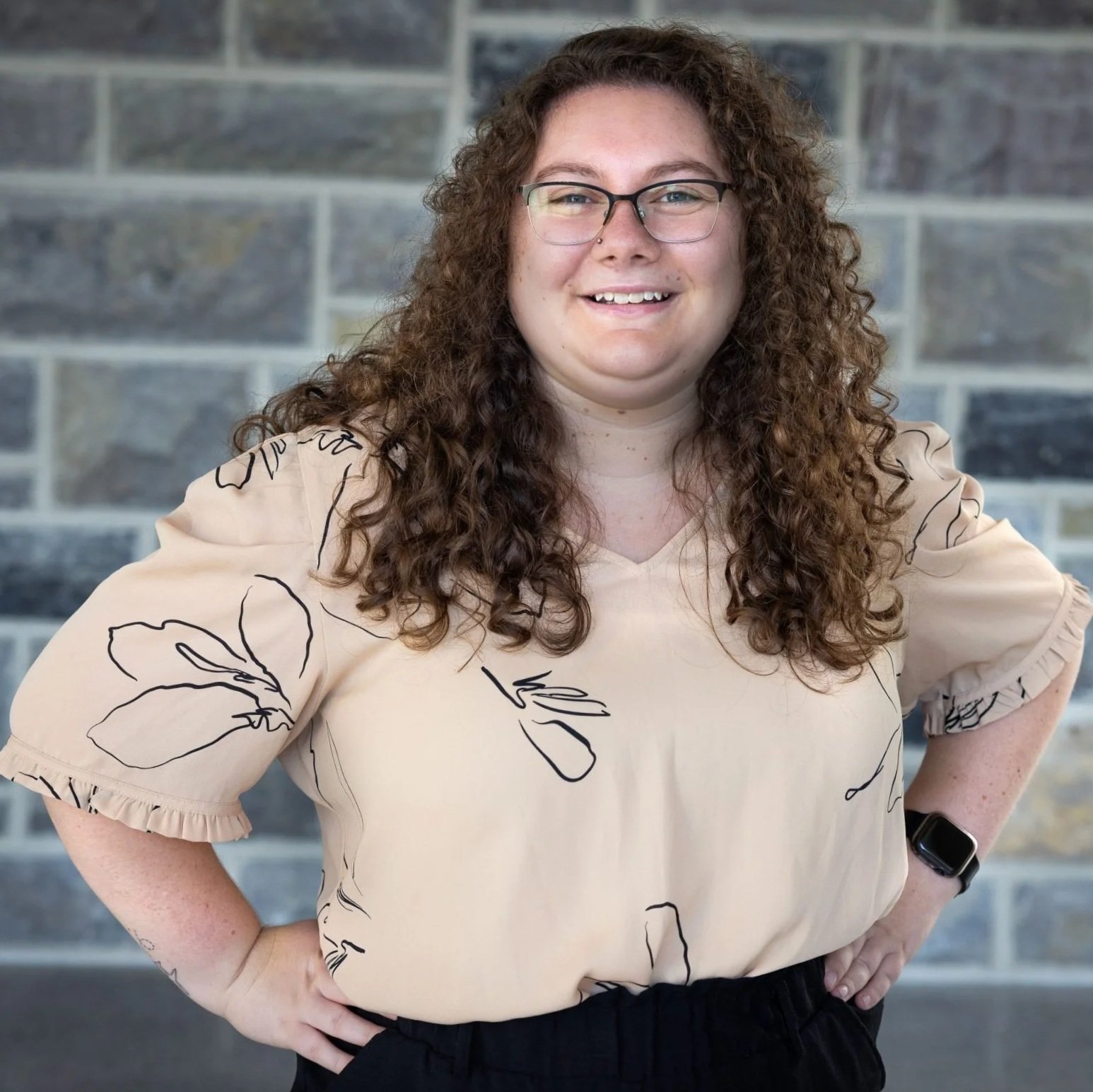 A woman with curly hair and glasses standing in front of a brick wall, wearing a beige top with black floral outline patterns, with her left hand on her hip and smiling.