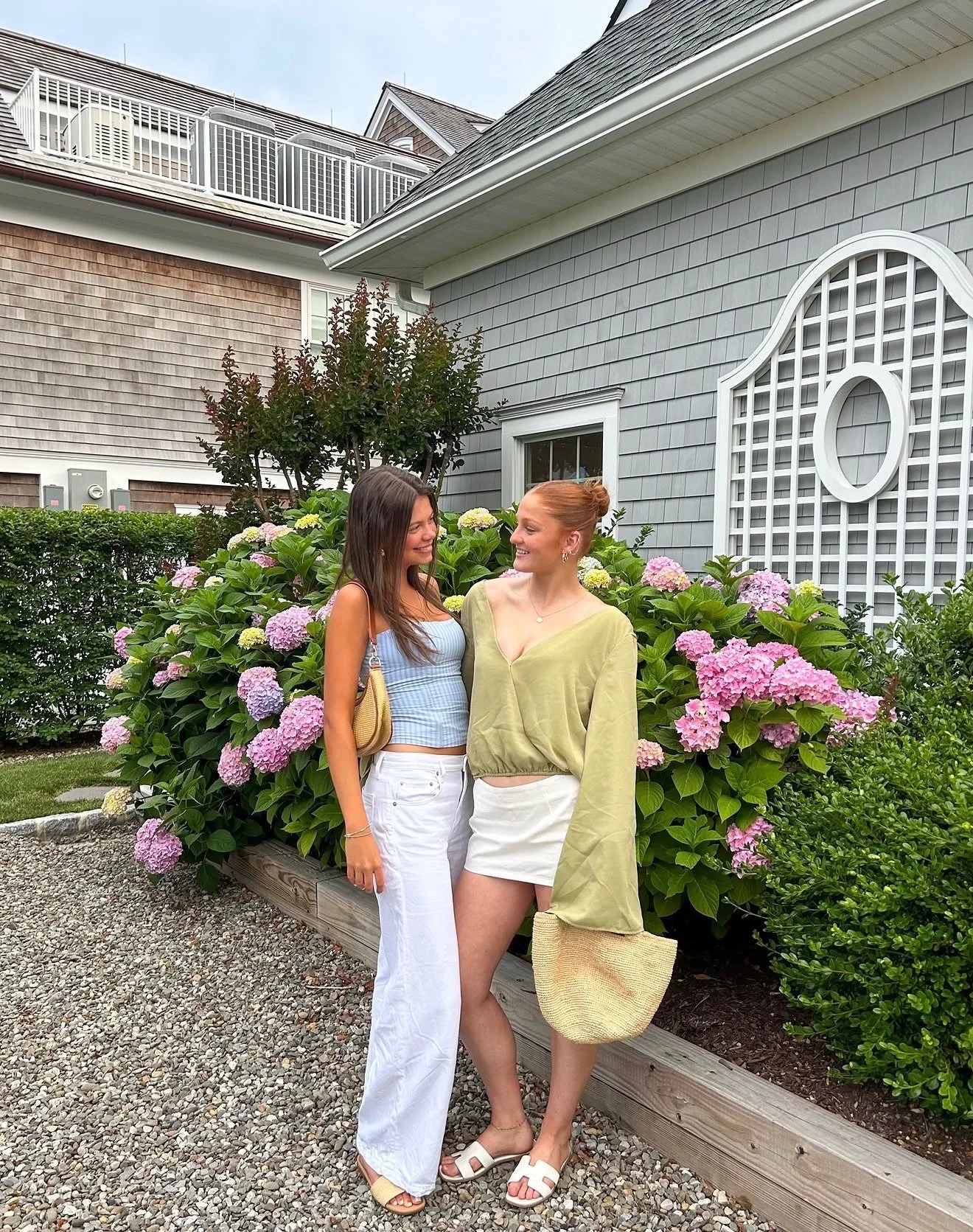 Two women standing in front of a bush with pink and purple hydrangeas, smiling and looking at each other, with a house and a gray fence in the background.