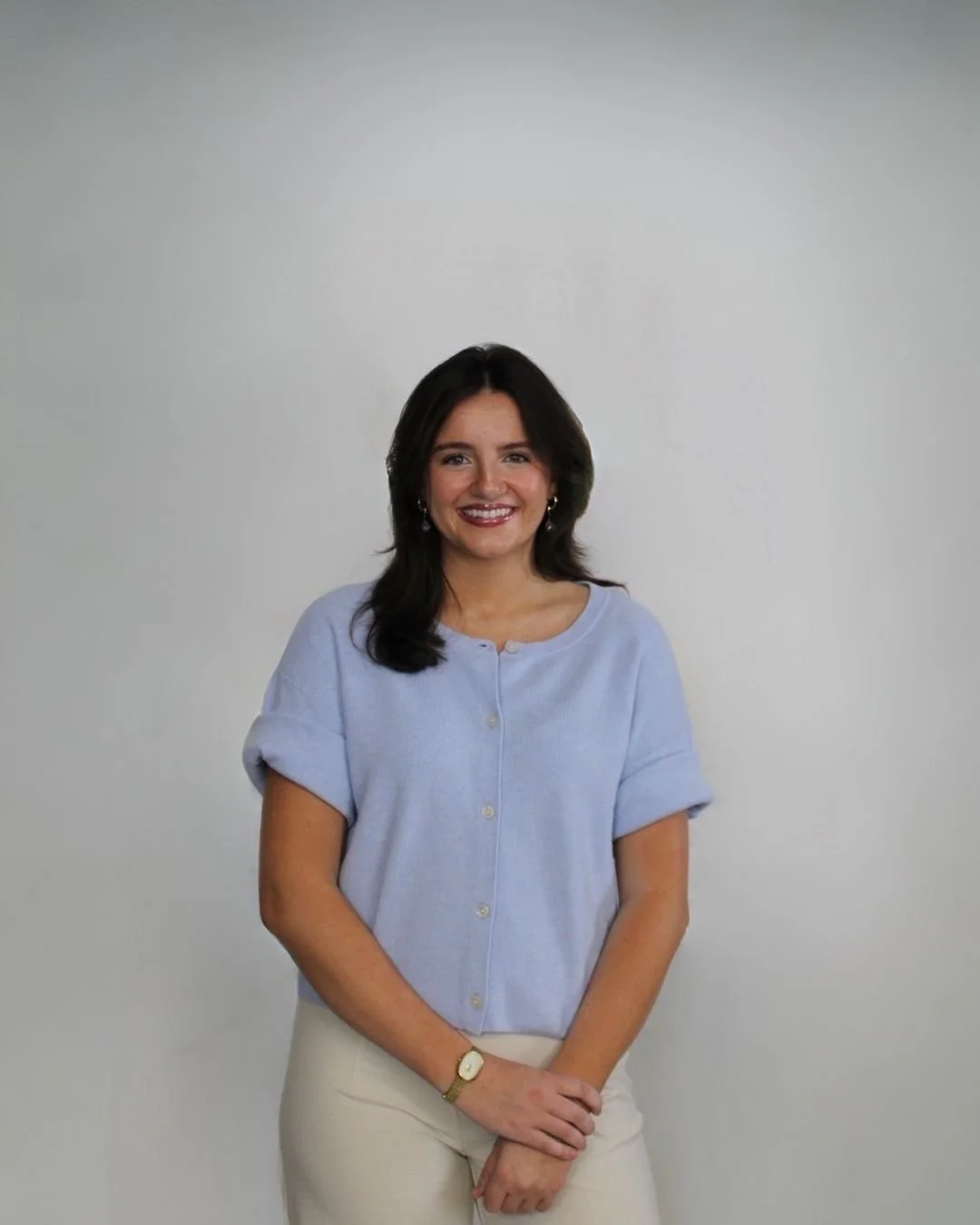 A woman with shoulder-length dark hair, smiling, wearing a light blue short-sleeved button-up shirt and beige pants, standing against a plain light background.