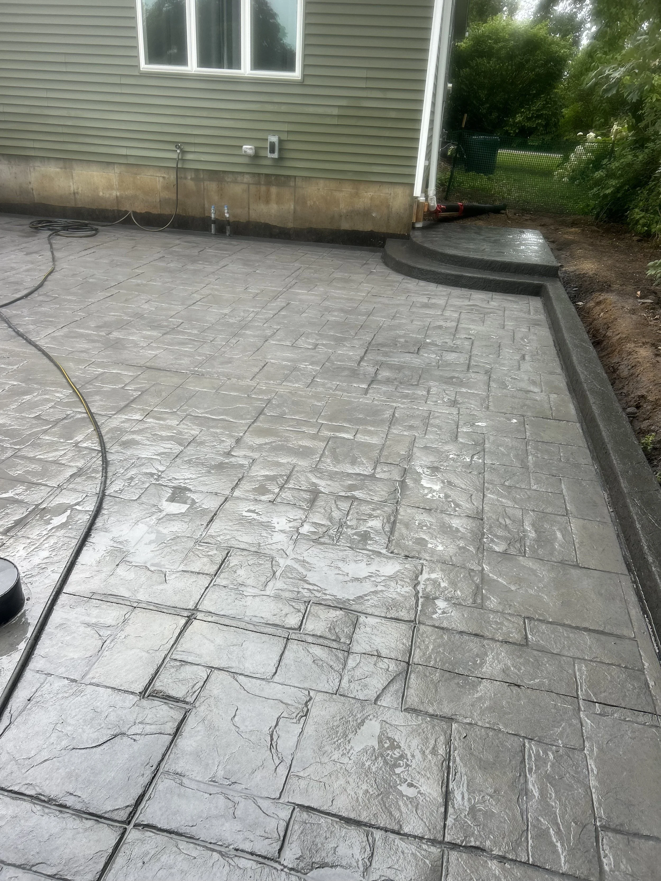 Backyard patio with newly installed gray stone concrete pavers, a curved step leading to a grassy yard surrounded by trees, a neighboring house with green siding, window, and utility hookups visible.