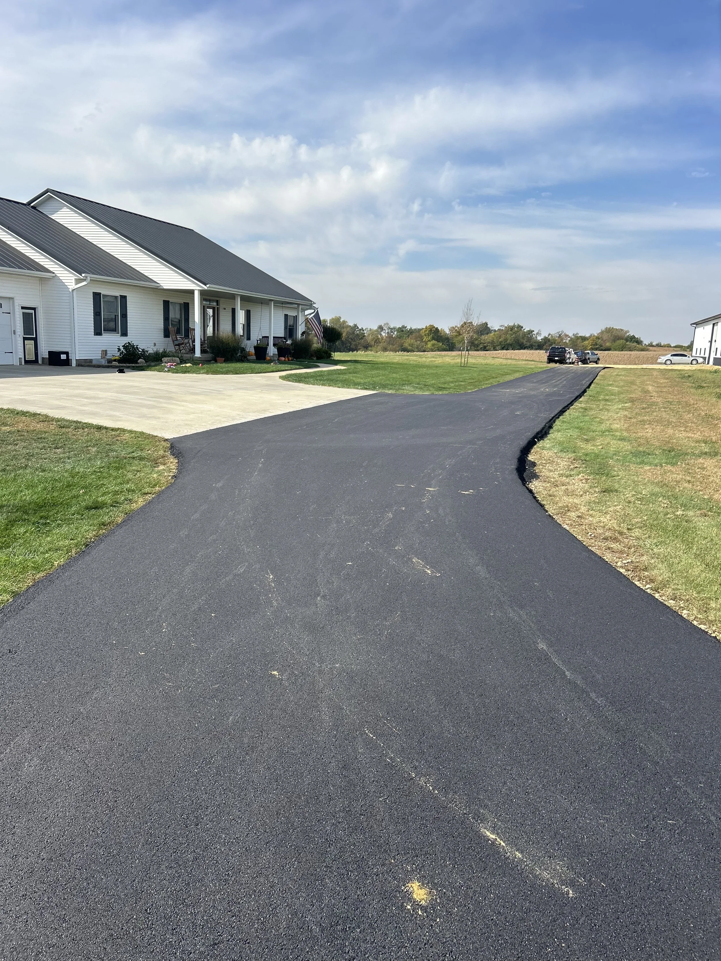 A newly paved black asphalt driveway leading to a house with a white exterior and dark shutters. There are several parked cars in the background, and a grassy lawn surrounds the driveway. The sky is partly cloudy with patches of blue.