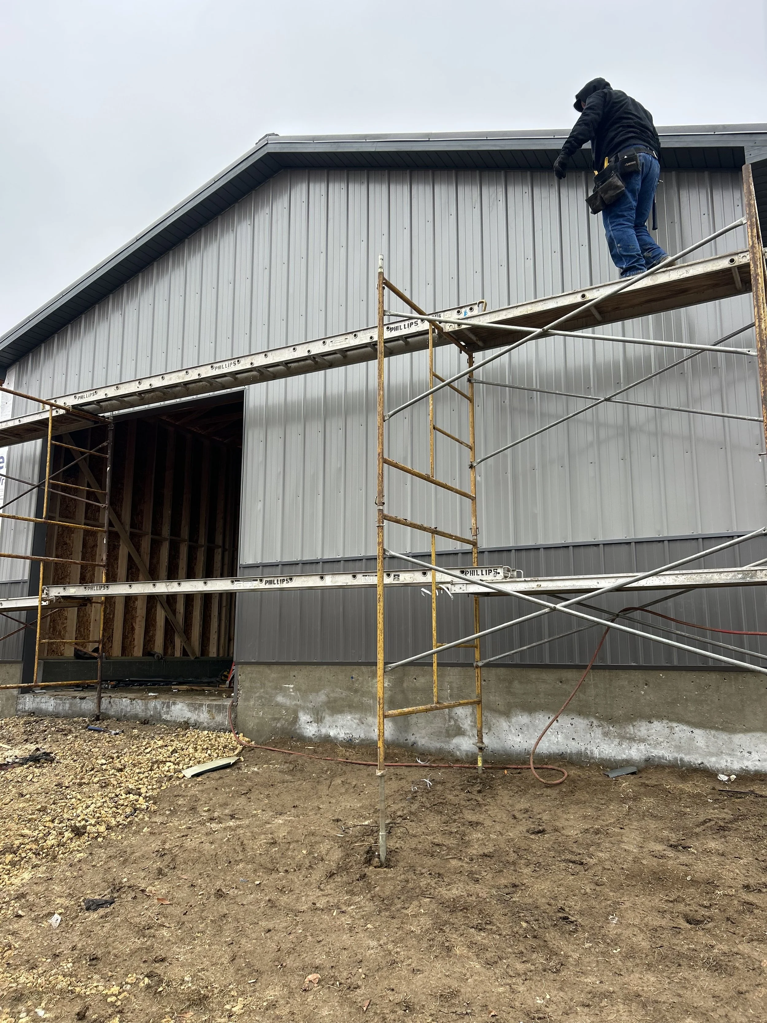 A construction worker standing on scaffolding installing or repairing the exterior metal siding of a building. The ground is bare dirt and the sky is overcast.
