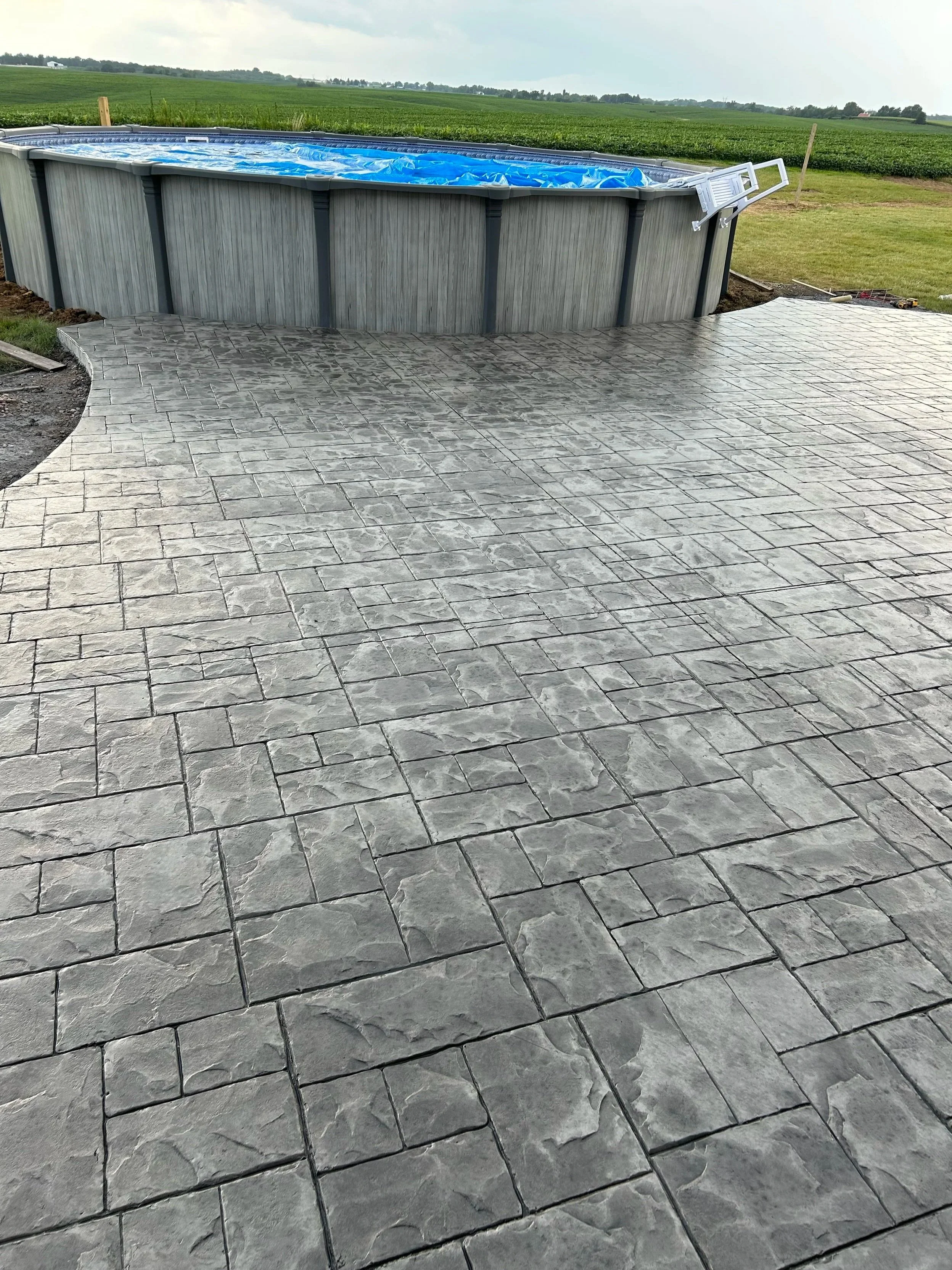 Above-ground oval swimming pool with a blue cover, situated next to a newly paved gray stone patio, with green fields and cloudy sky in the background.