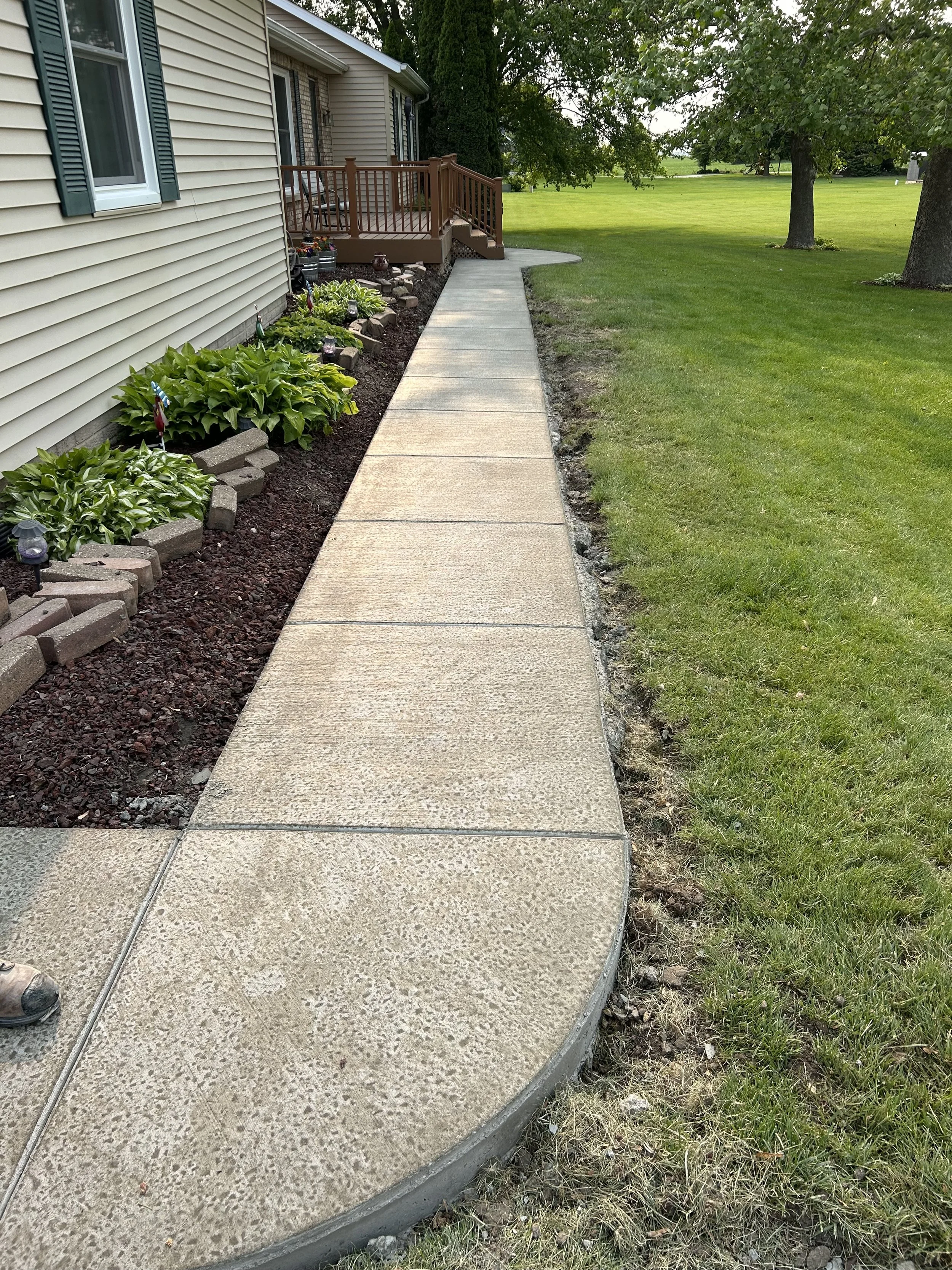 A concrete sidewalk next to a house with beige siding and green shutters, with a green lawn and trees in the background.