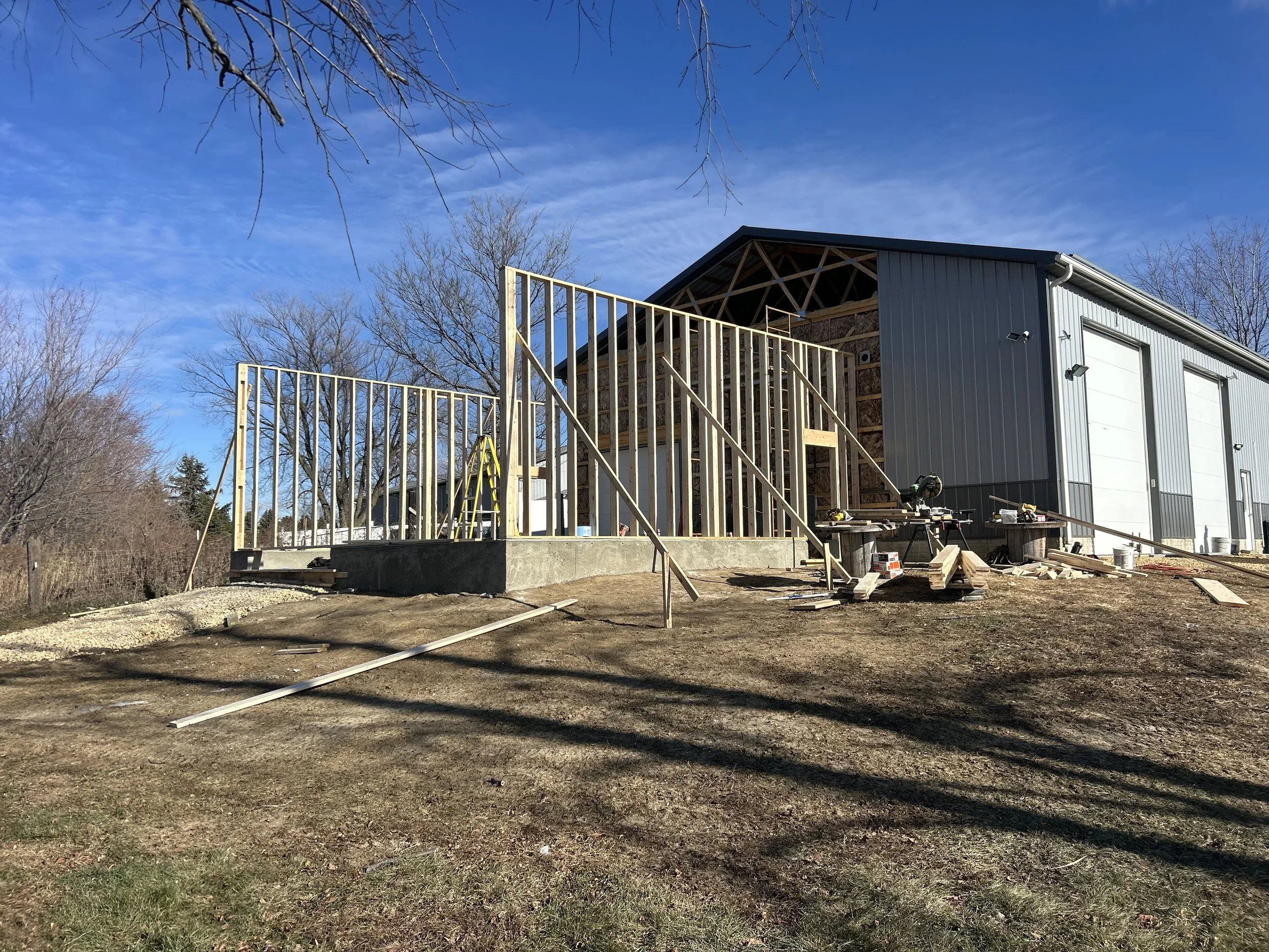 Construction site with a building frame under construction, partially covered with metal siding, and wooden framework for additional structure, with blue sky and leafless trees in the background.