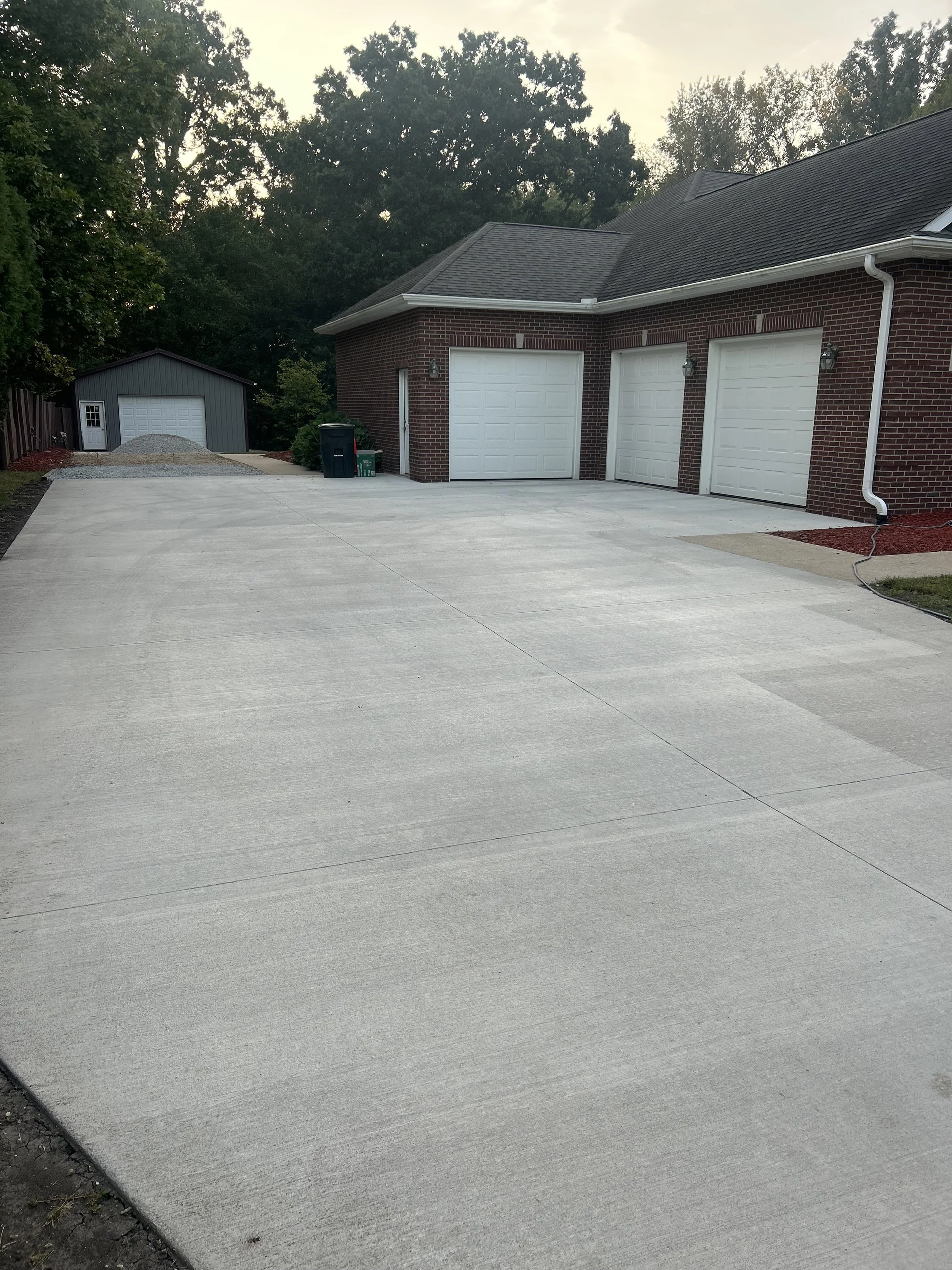 A newly paved concrete driveway in front of a brick garage with two doors, surrounded by some trees and a small shed in the background.