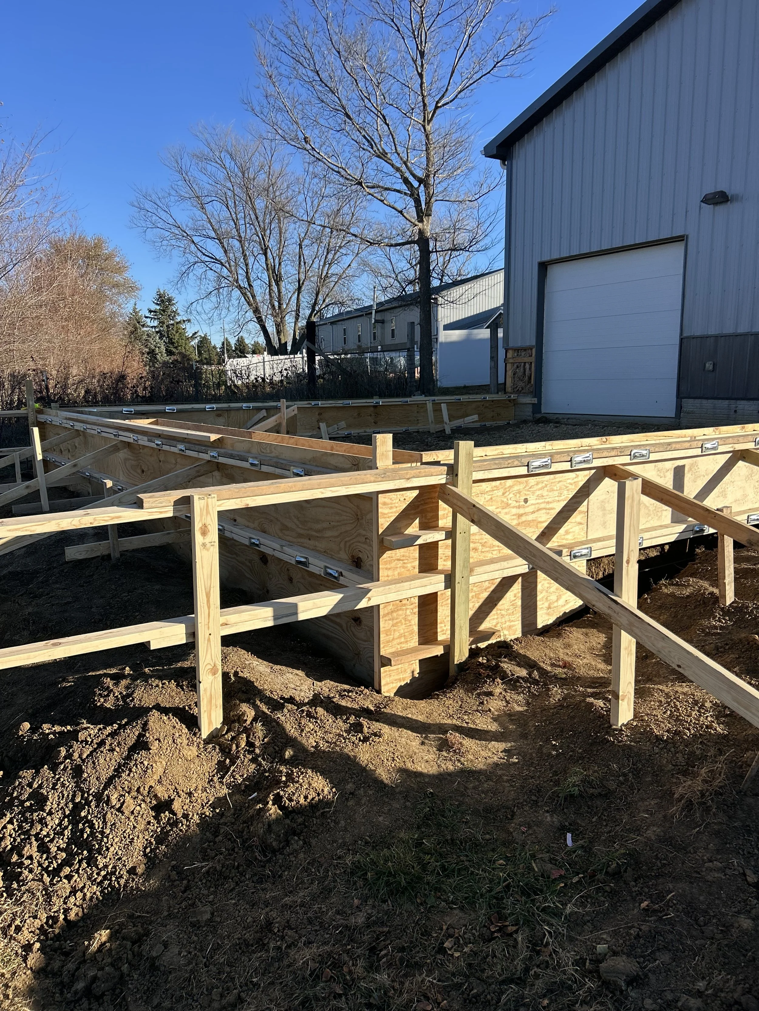 Wooden framing for a concrete slab foundation in progress outside a building, with a large tree and clear blue sky in the background.